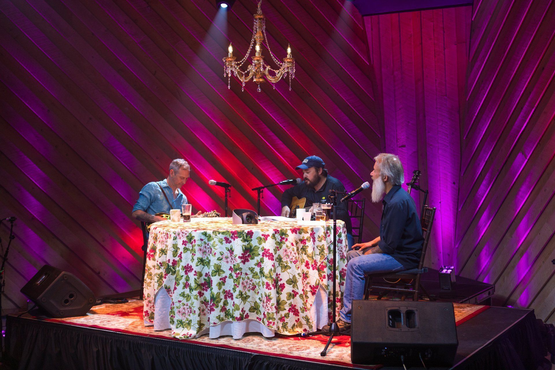 Three people seated at microphones around a floral table on a stage with pink drapes and a chandelier.