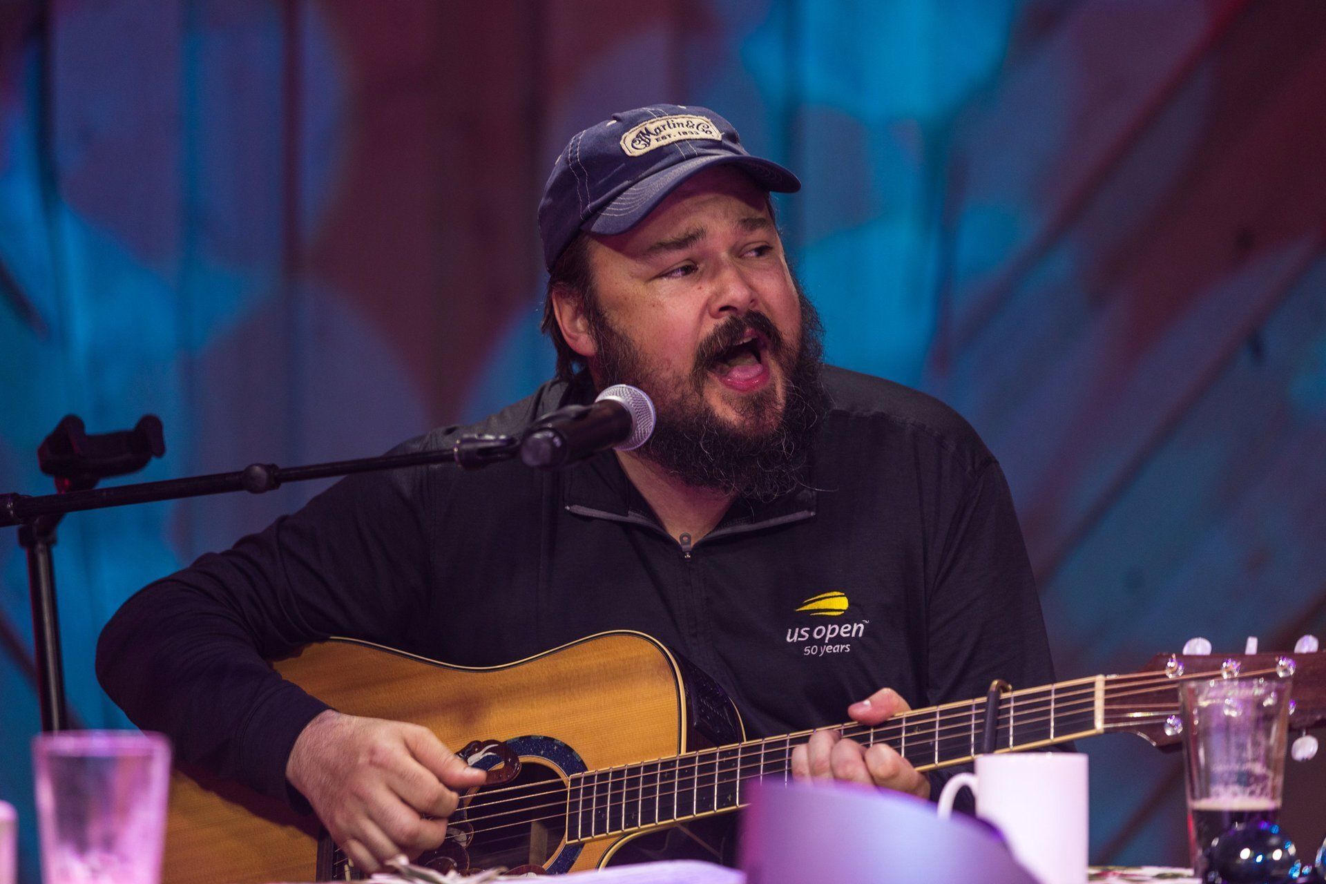 Musician singing into a microphone while playing acoustic guitar onstage in a black shirt and cap