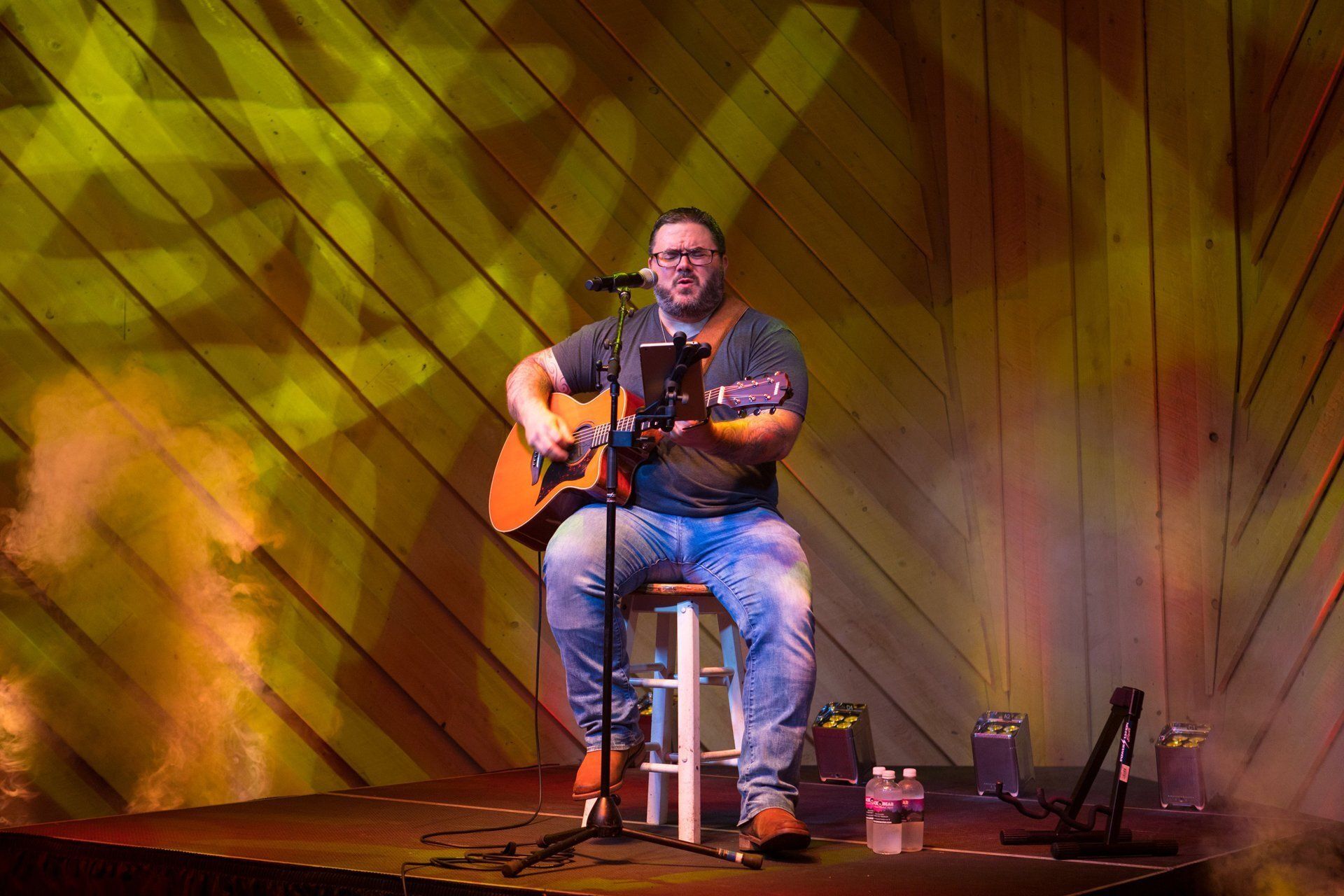 Musician seated on a stool, playing guitar on a warmly lit stage with a wooden backdrop.
