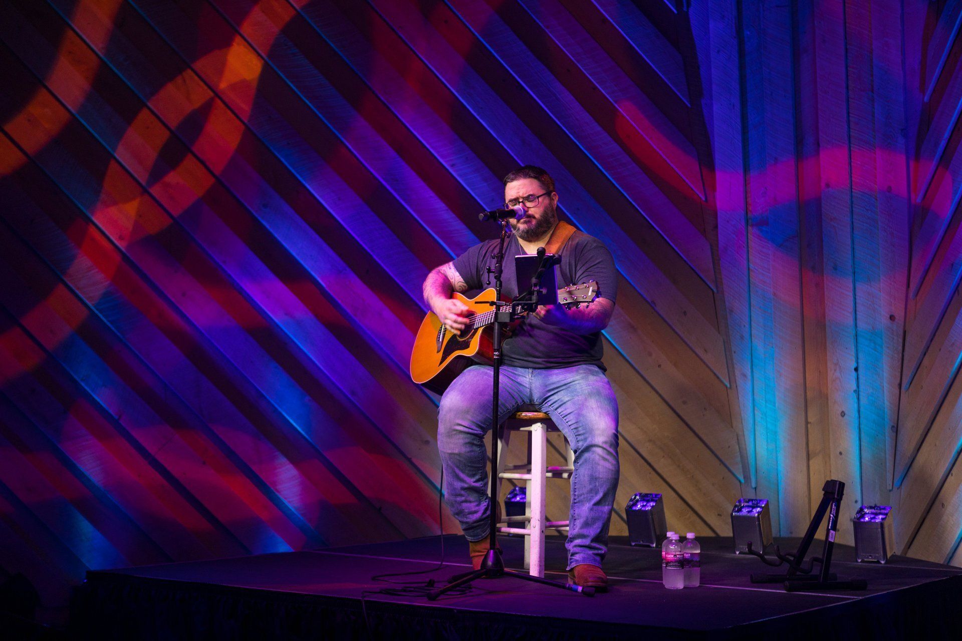 Musician seated onstage, playing acoustic guitar under purple and blue lights