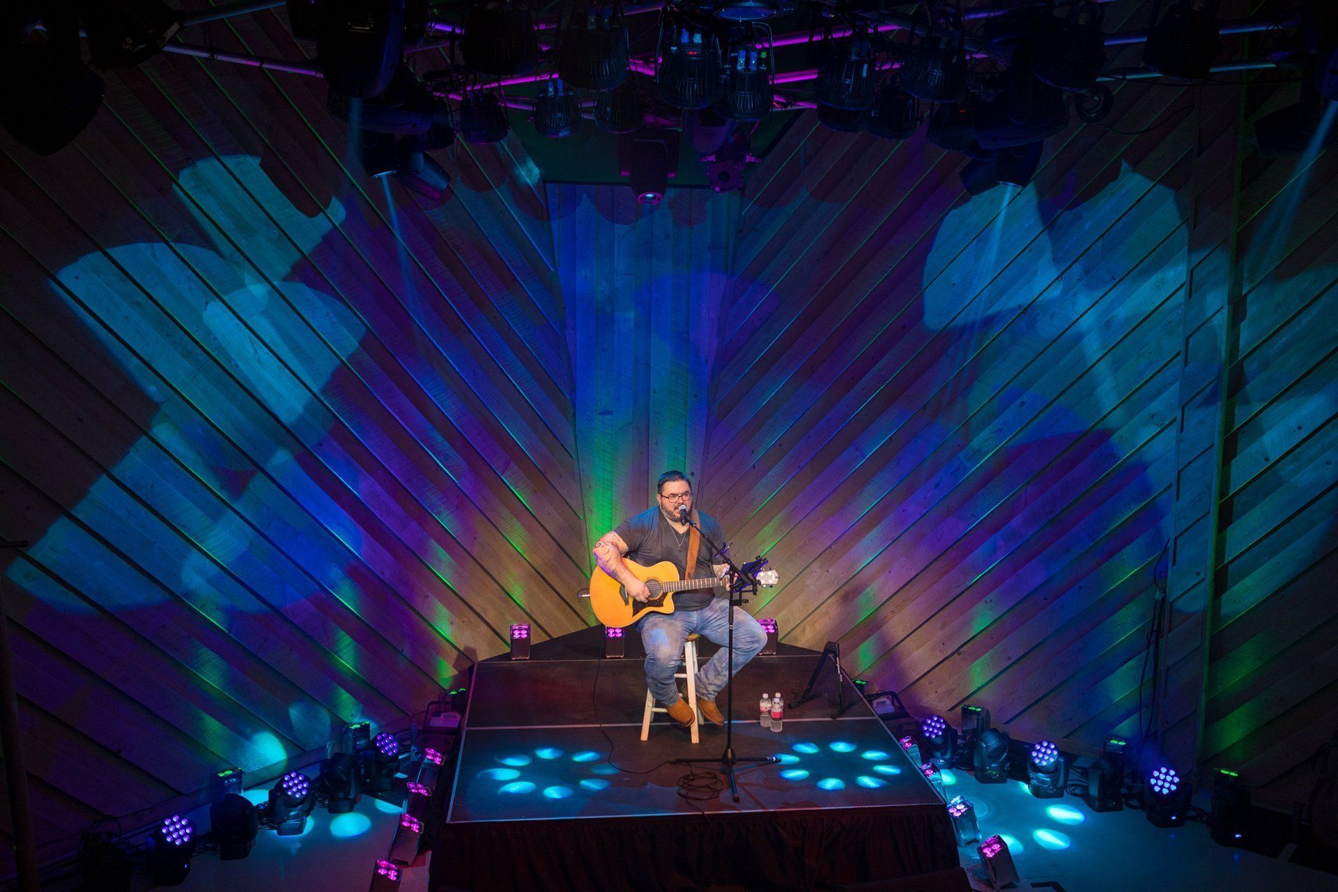 Musician performing on a lit stage with guitar, blue and purple lights behind him.