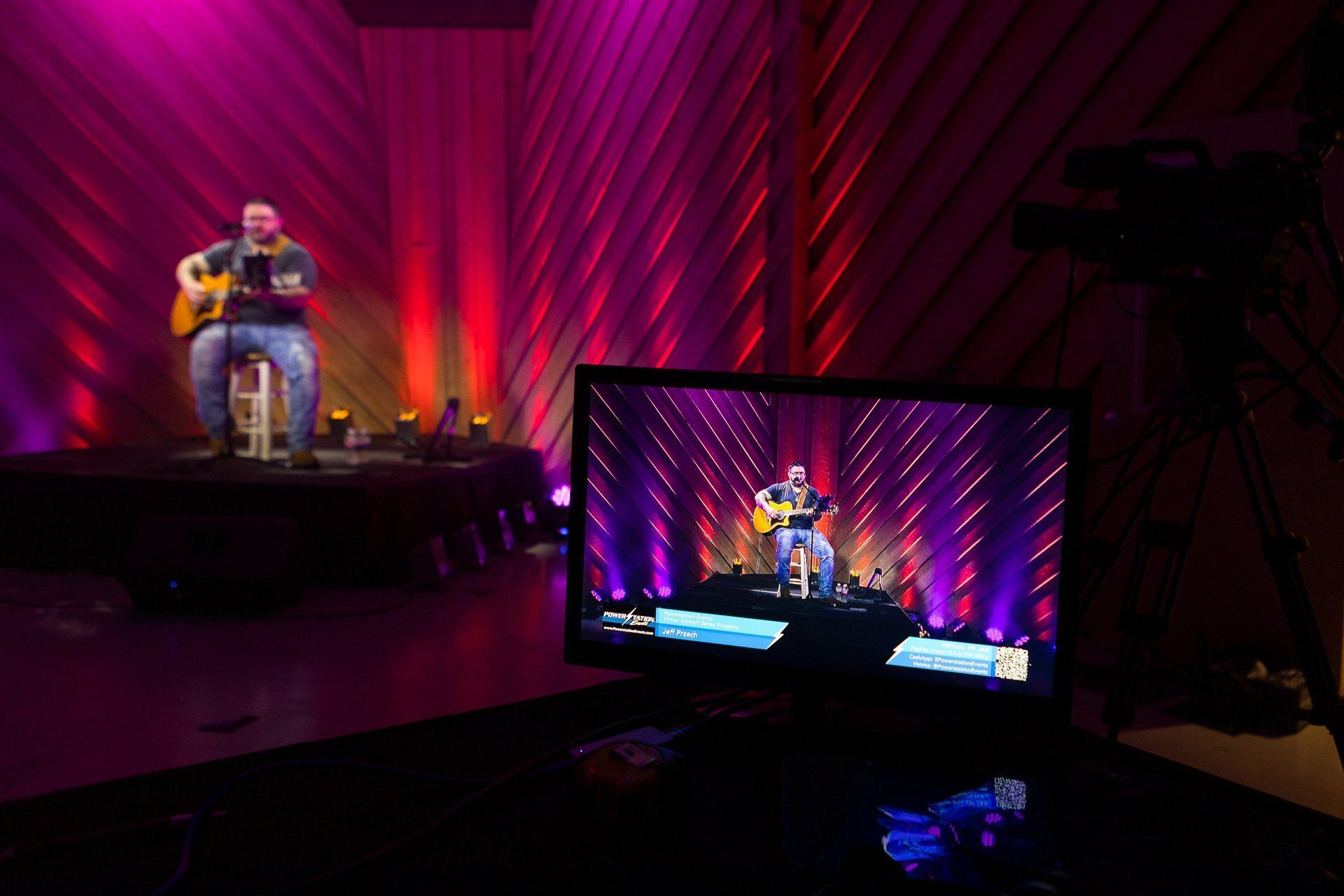 Musician on a small stage with purple-red lighting, seen on a monitor in a dark studio