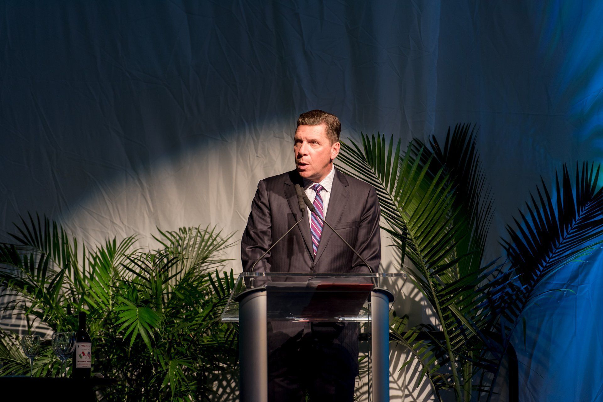 Man speaking at a podium on a stage with palm plants and blue lighting