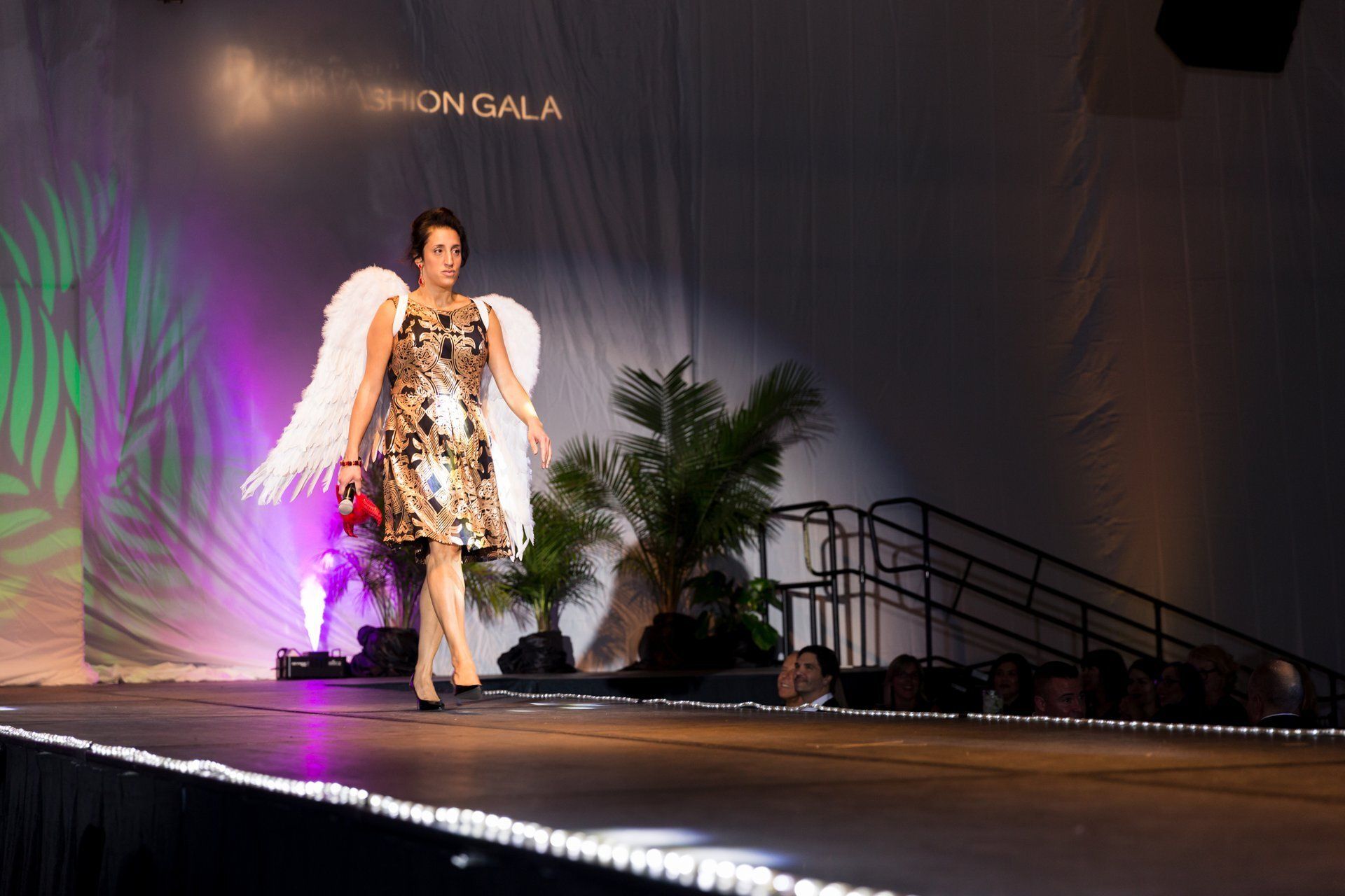 Winged model in a patterned dress on a lit fashion runway, with stairs and purple stage lighting.