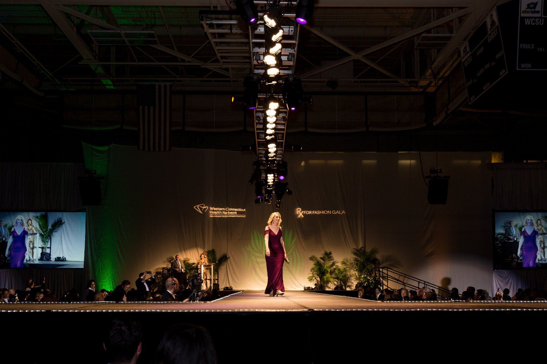 Fashion runway with a model in a burgundy dress under bright overhead lights, flanked by plants and a screen display.