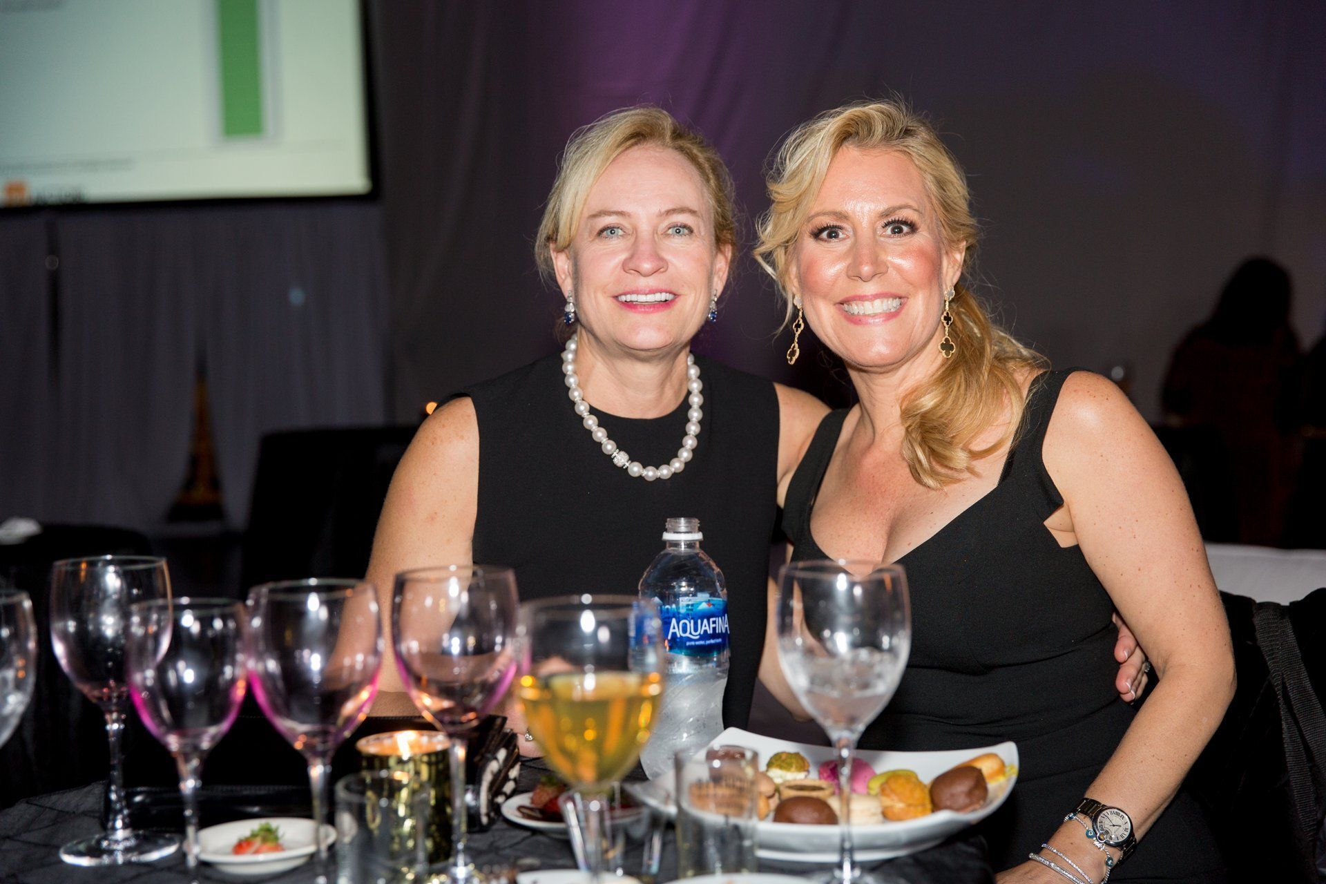 Two people seated at a dinner table, smiling at the camera with wine glasses and plates of food.