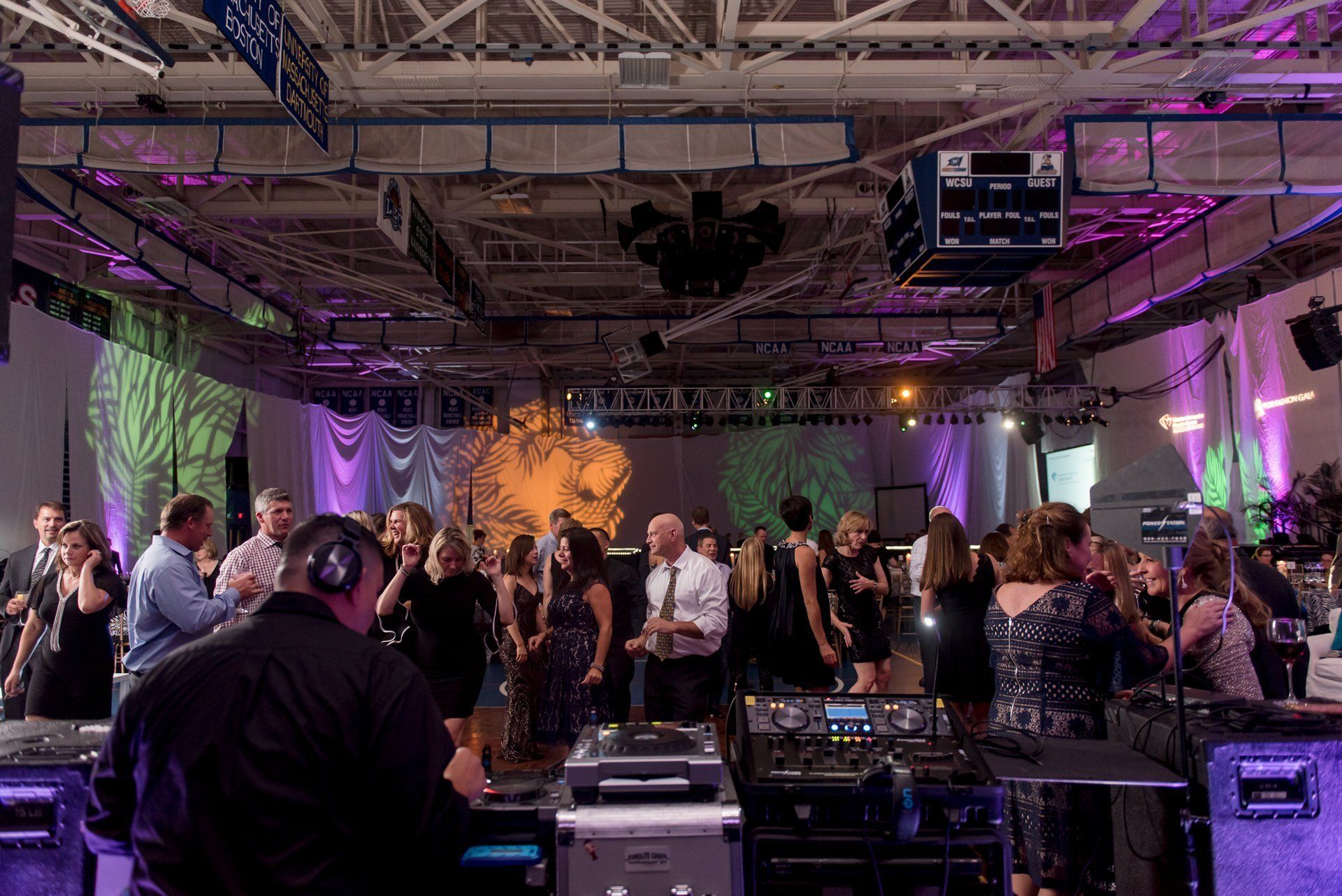 People dancing at a party in a large hall with purple stage lighting and sound equipment in front.