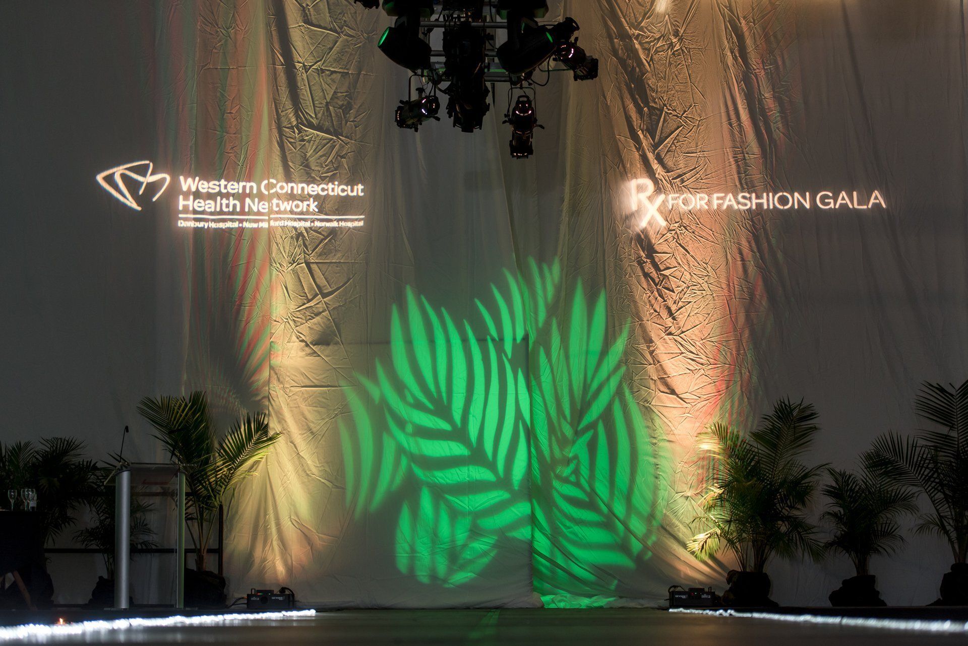 Fashion gala stage with green leaf projection, plants, lights, and event logos on a draped backdrop