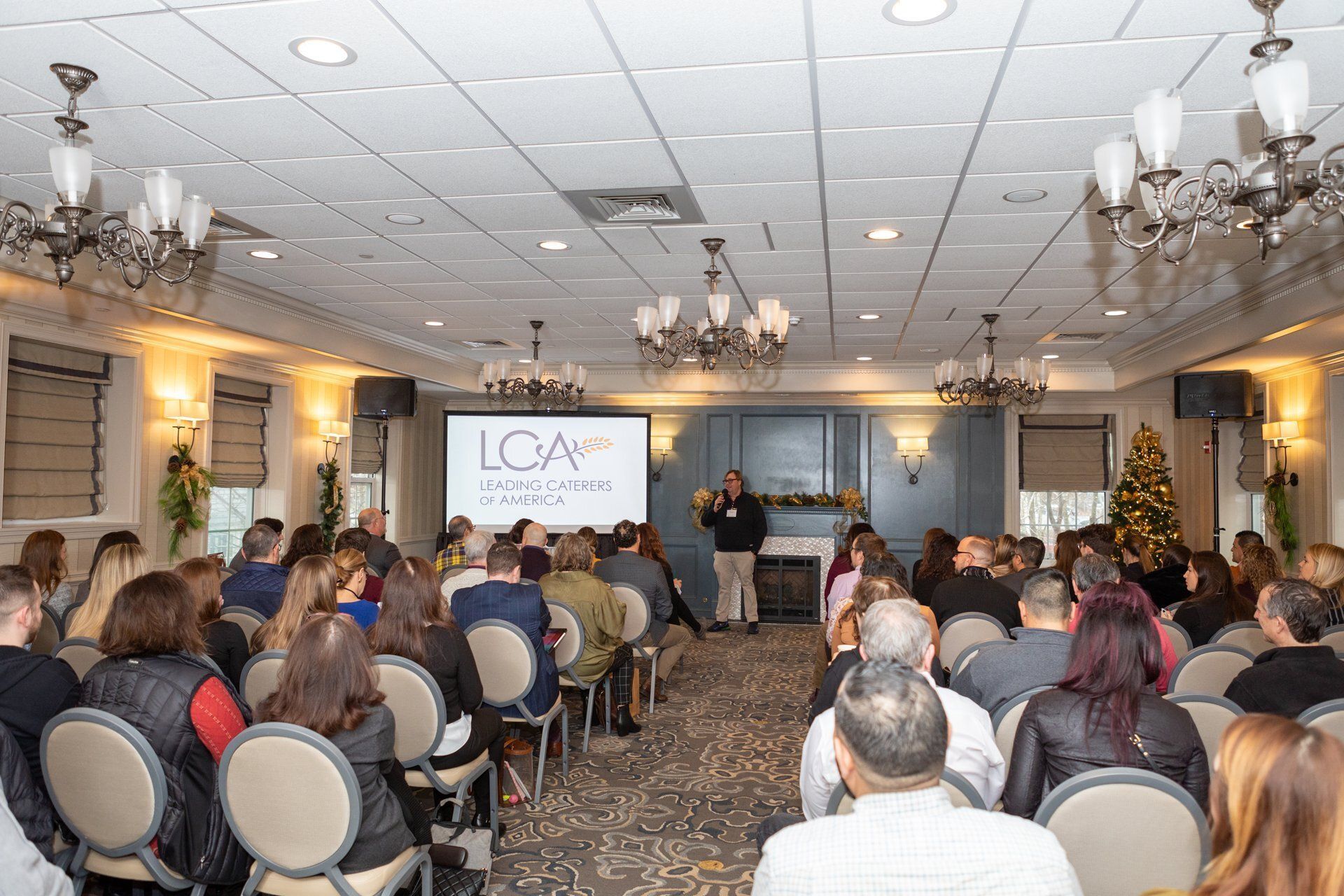 Conference audience facing a speaker at the front of a decorated banquet hall