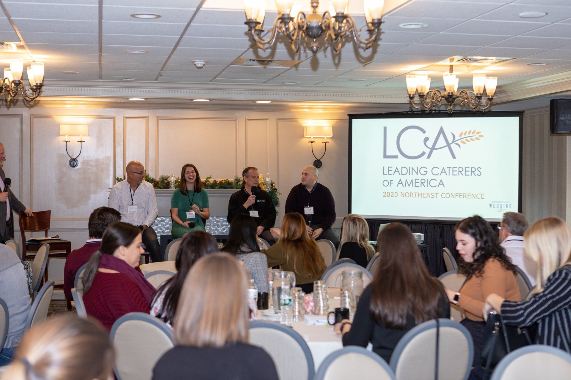 Panel discussion at a conference luncheon, with speakers on stage and audience seated at round tables