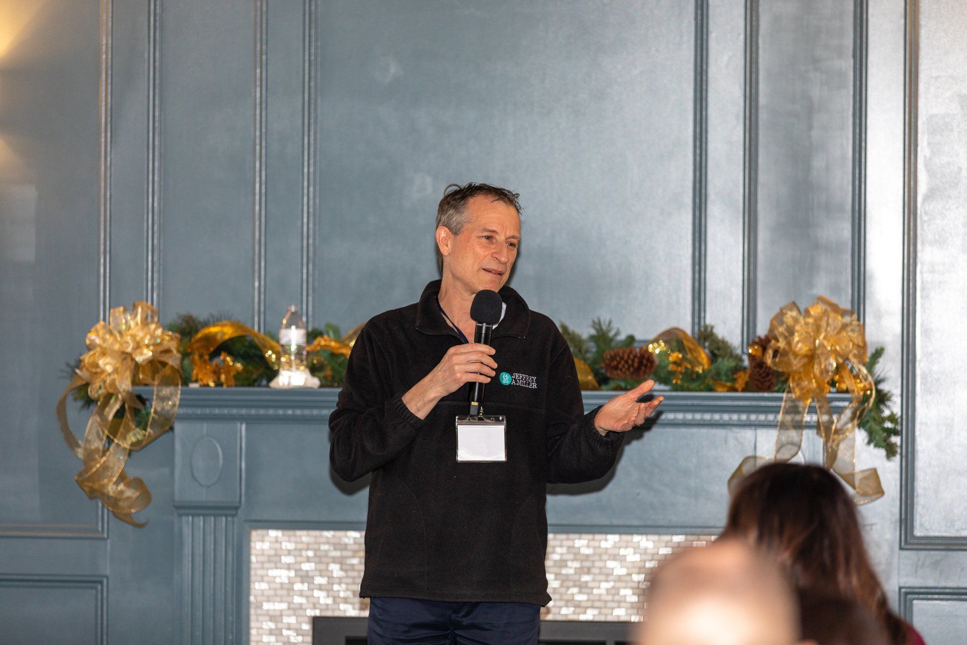 Person speaking into a microphone on a decorated stage, holding a remote, with holiday garlands behind them.