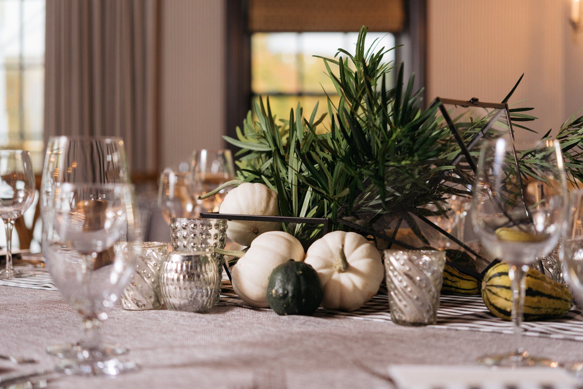 Elegant table setting with white pumpkins, candles, glassware, and greenery on a white tablecloth