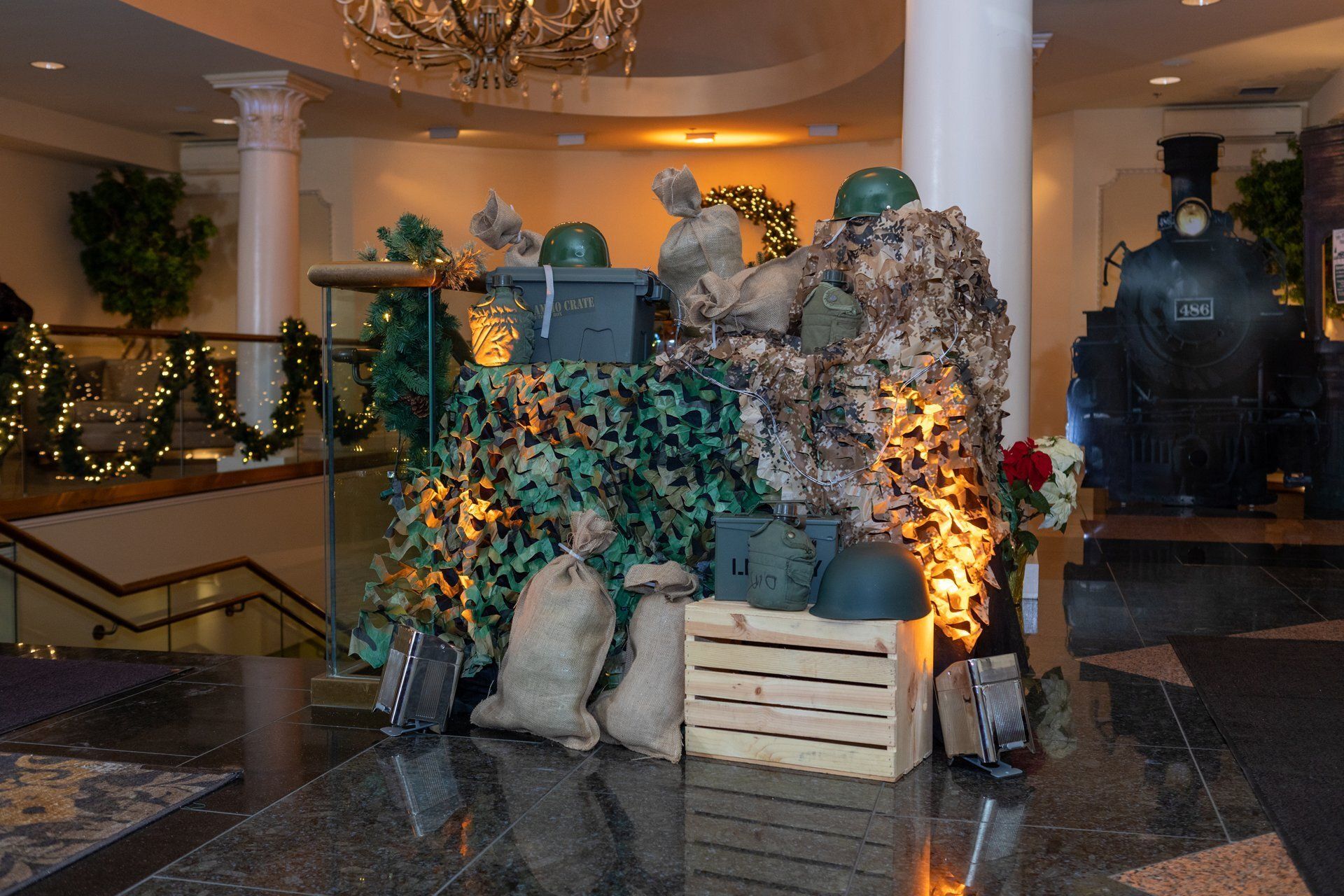 Indoor holiday display with stacked crates, faux rocks, and greenery lit by warm orange lights in a lobby