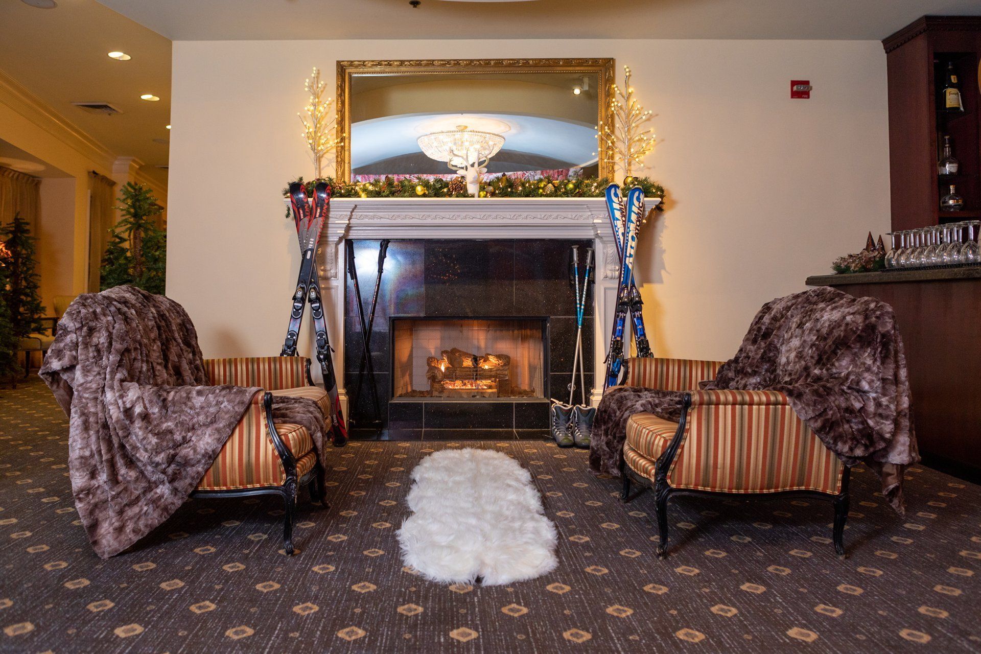 Elegant lobby with a lit fireplace, mirrored mantel, two striped chairs, and fur throws on a patterned carpet