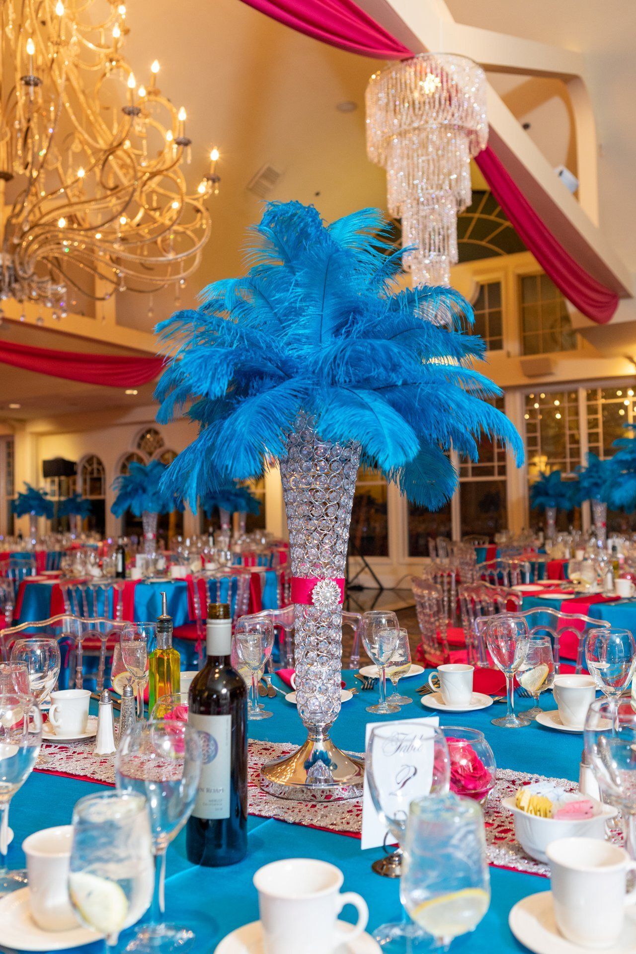 Elegant banquet hall with blue feather centerpieces, white tables, and pink drapery under chandeliers
