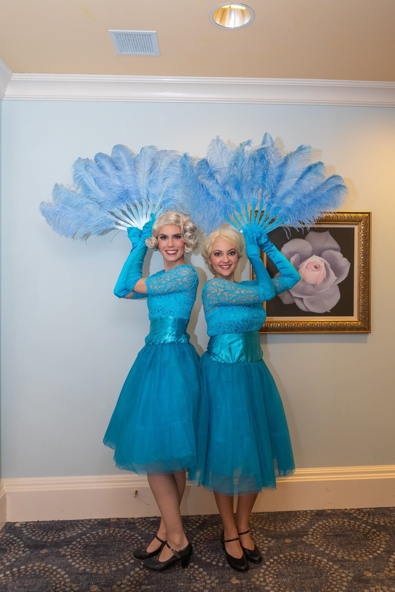 Two performers in turquoise dresses and feather fans posing in a hallway beside a framed painting