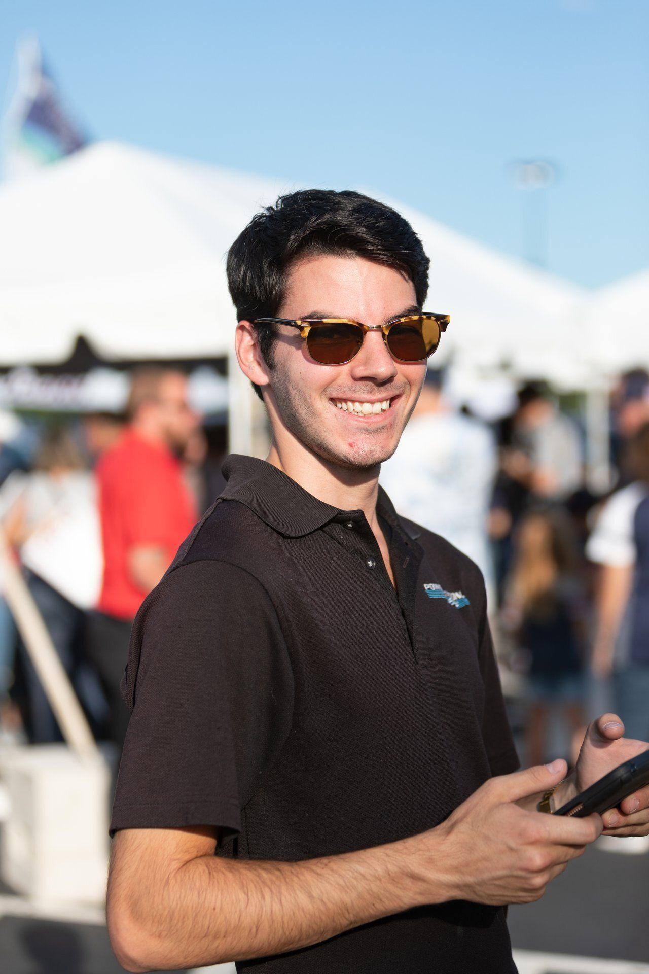 Smiling man in sunglasses holding a phone outdoors at a crowded event