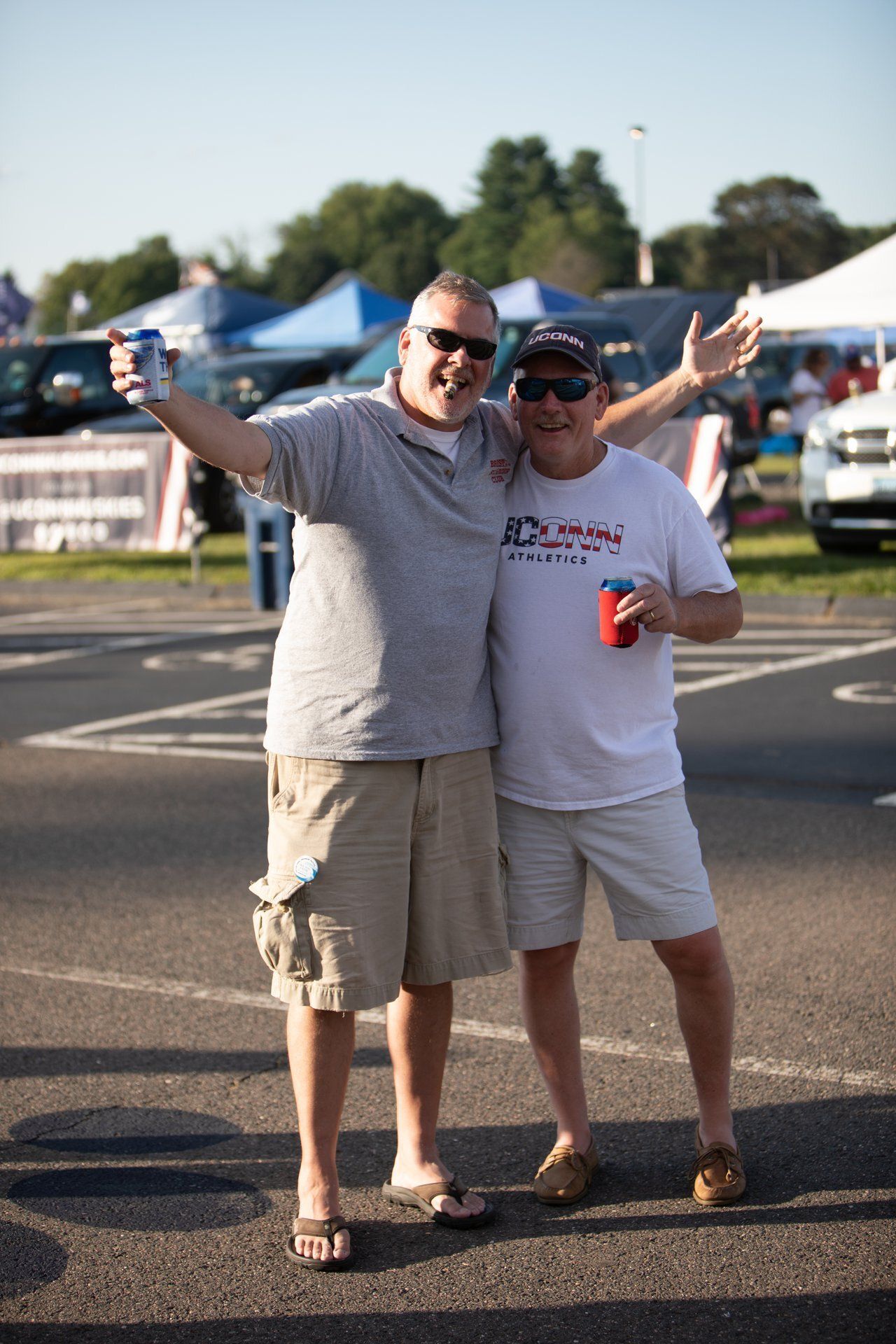 Two men posing together outdoors, one with arms raised, in a sunny parking lot near tents.