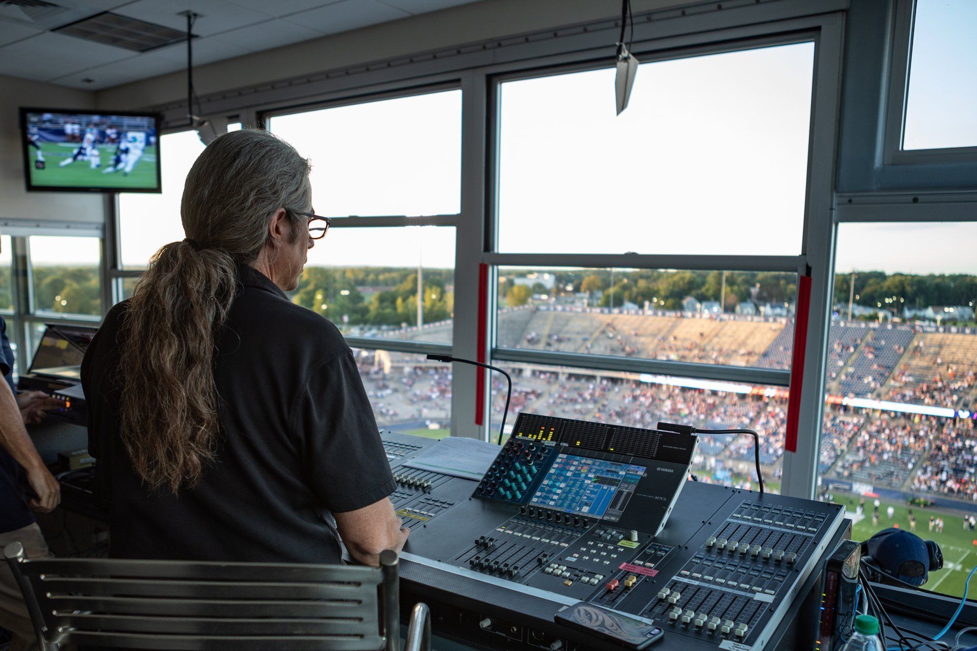 Person operating a sound mixing console in a stadium booth overlooking a live event