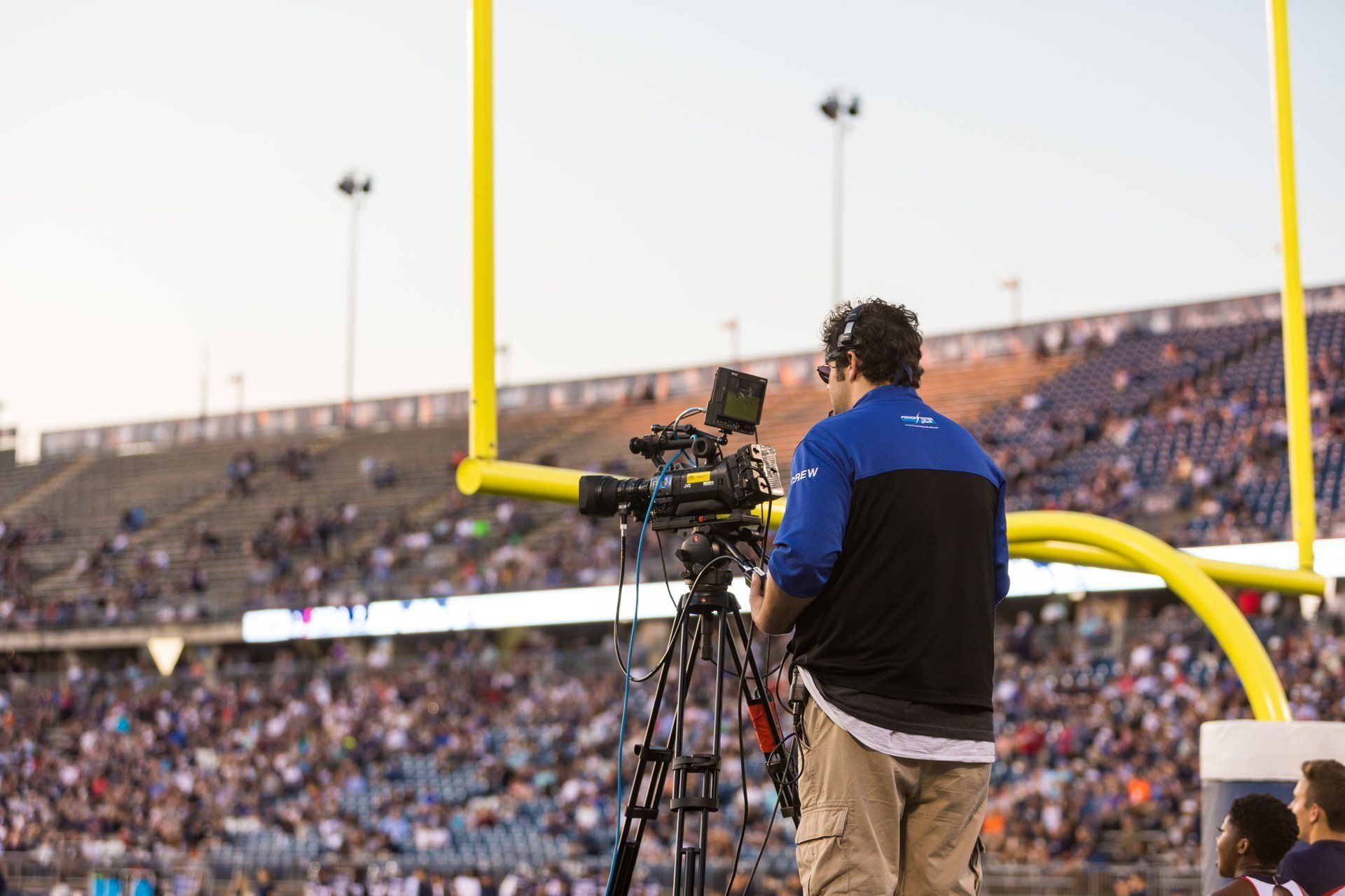 Camera operator filming a football game beside the goalpost, with stadium crowd in the background