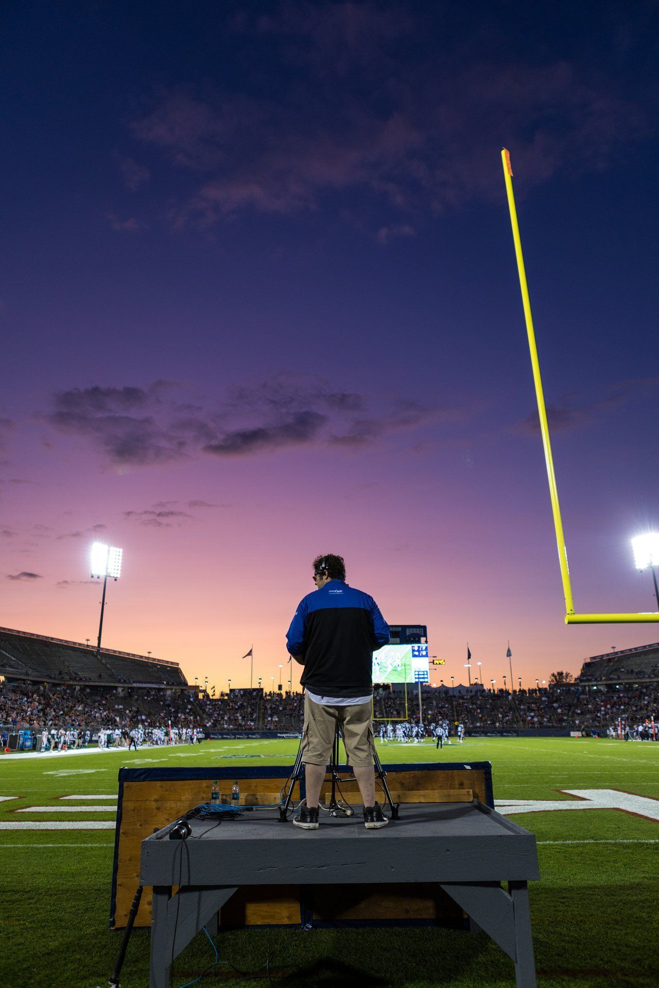 Person standing on a platform at a football stadium at dusk with a glowing yellow goalpost and crowd in the background