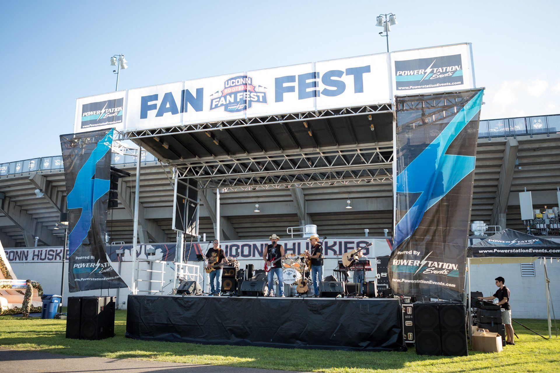 Outdoor FAN FEST stage with banners, speakers, and a few people performing on a sports field