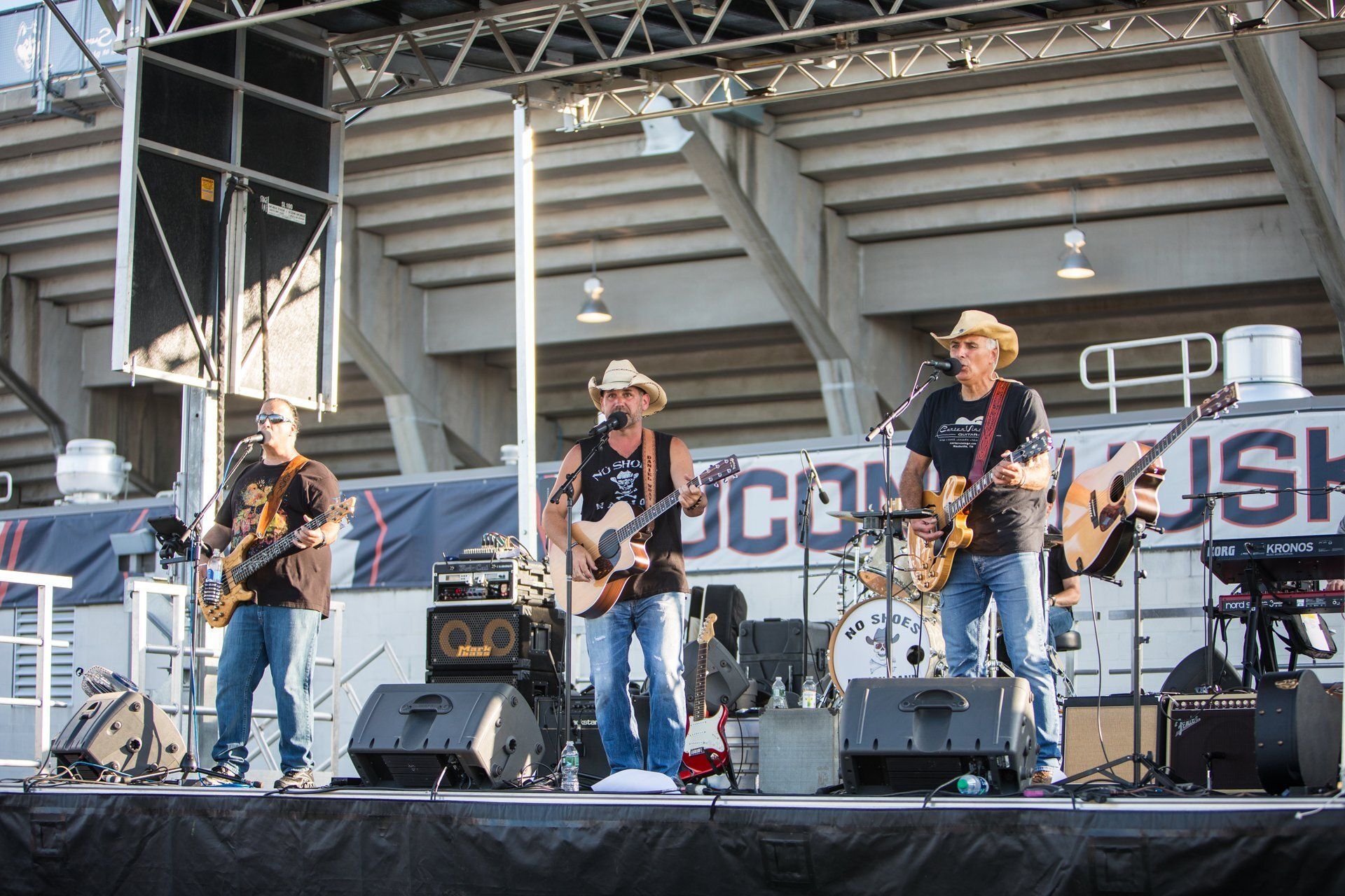 Country band performing on an outdoor stage with guitars and drums during daylight