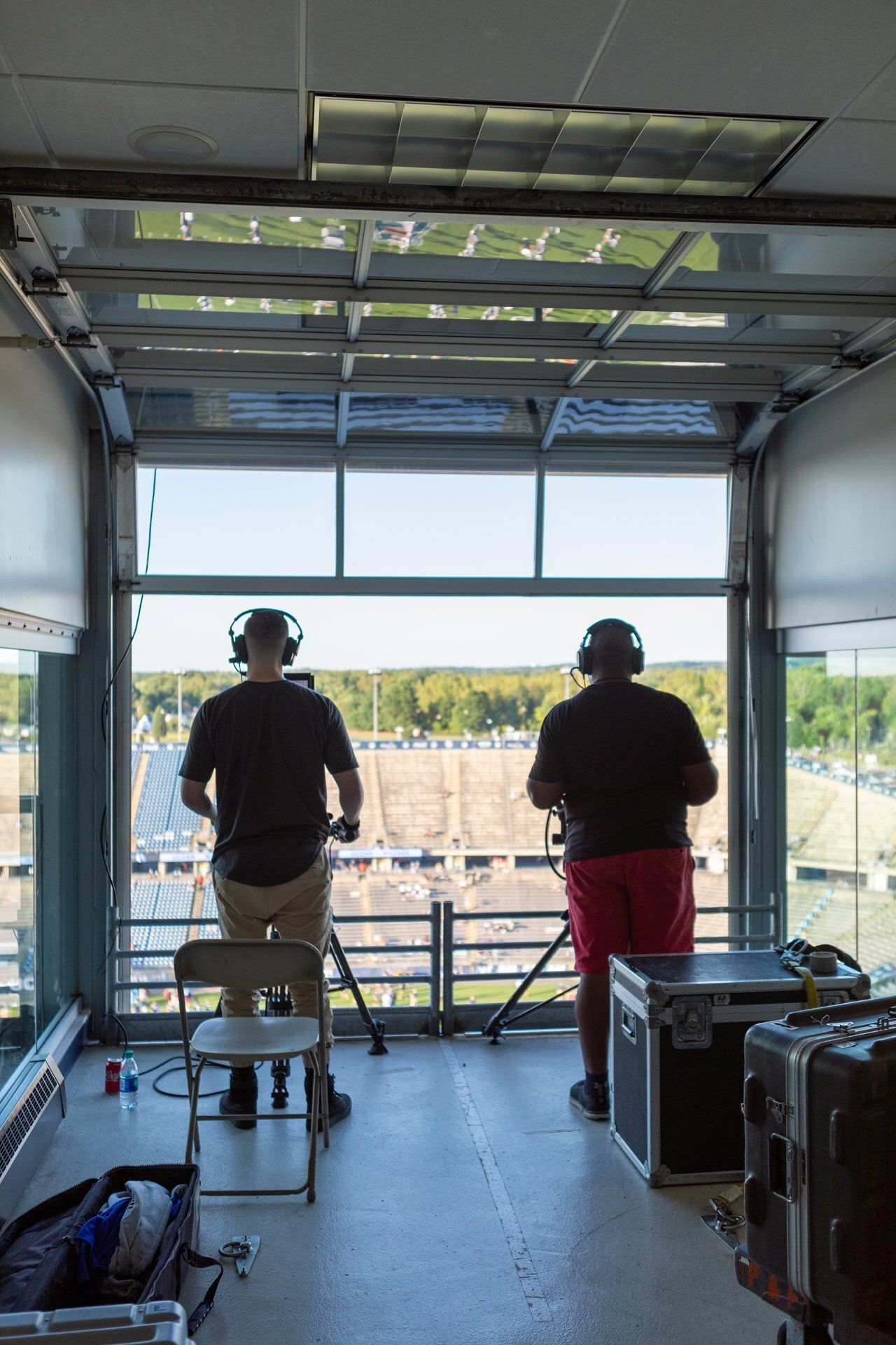 Two people stand by a window in a small room overlooking an outdoor area with equipment cases nearby.