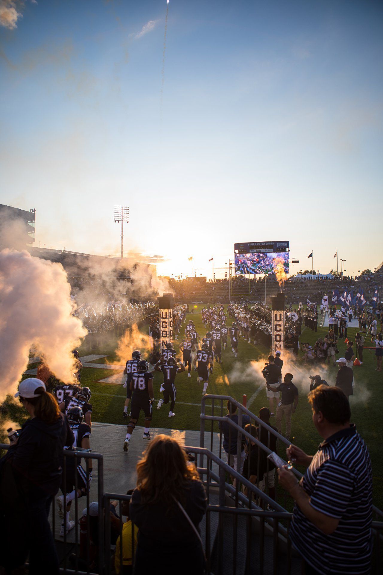 Crowded stadium during a smoky evening concert, with fans standing by railings and a lit stage in the distance