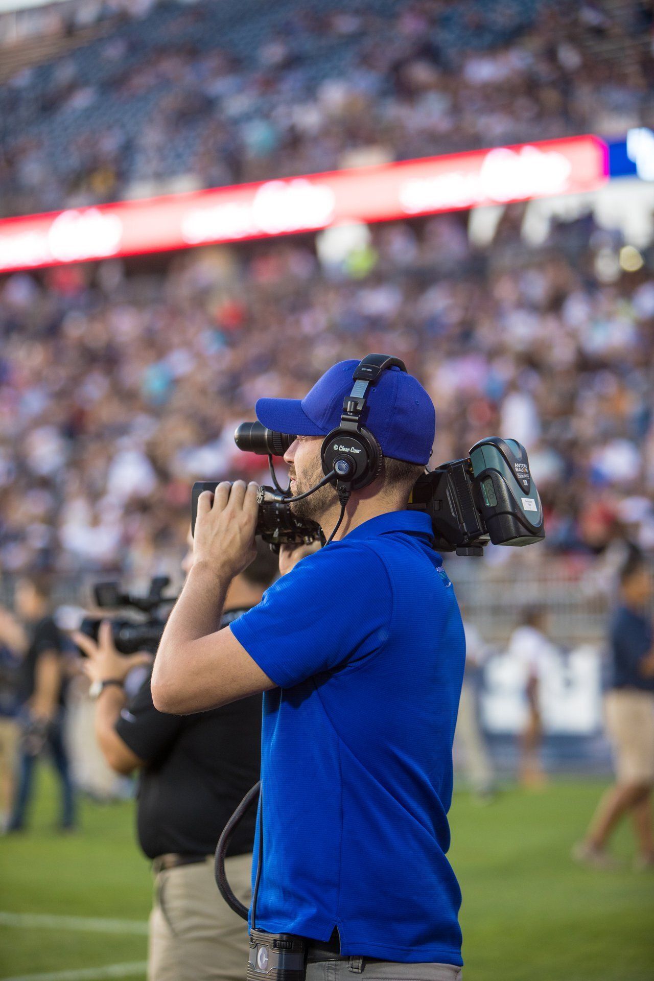Cameraman in blue shirt and cap filming on a football sideline, with a crowd blurred in the stadium background.