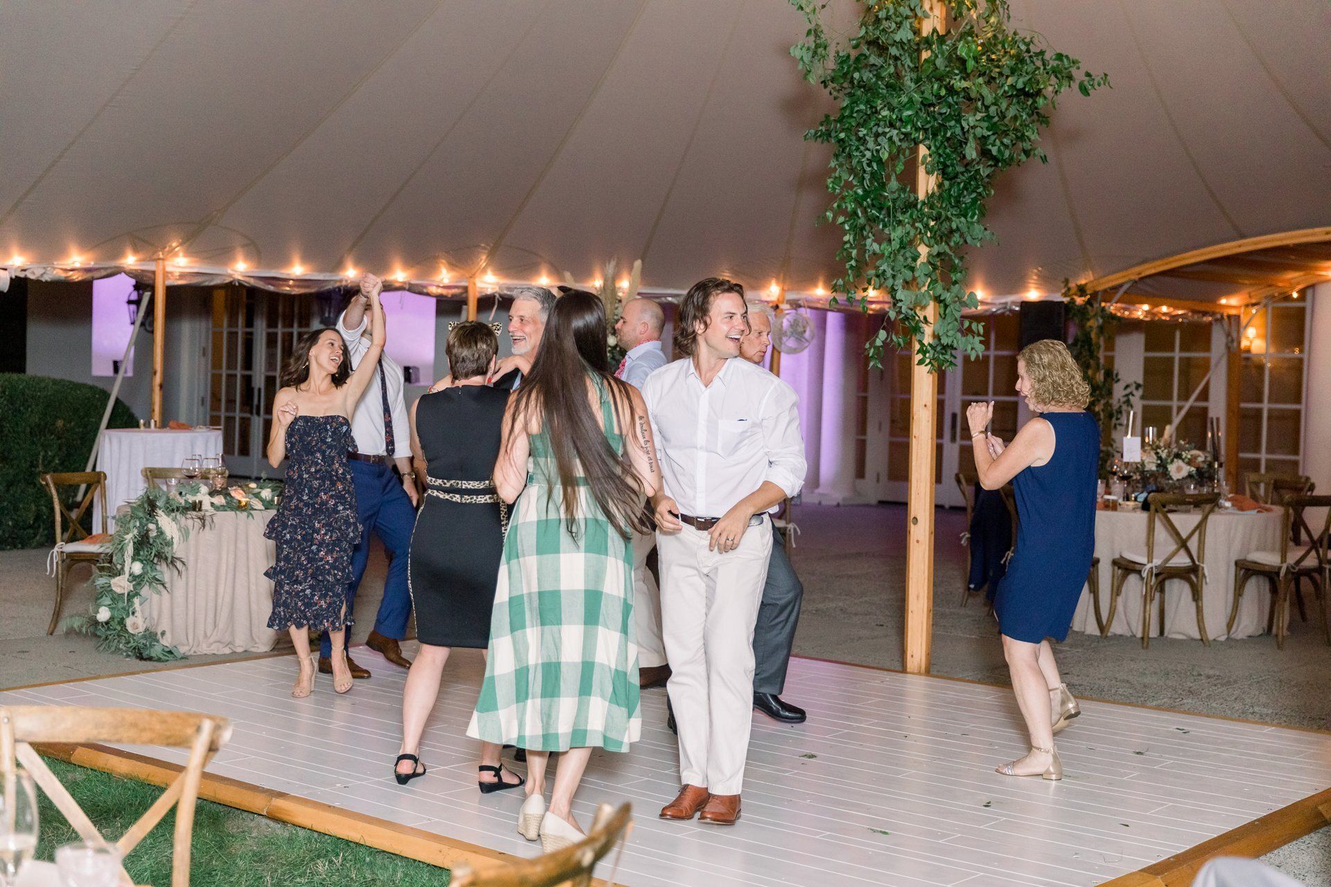 People dancing at an indoor event in a tented venue with string lights and greenery.
