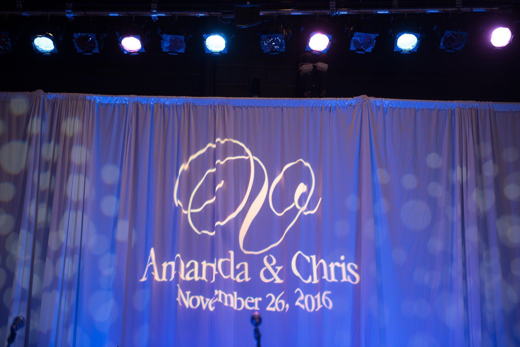 Projected wedding monogram and names “Anand & Chris” with date “November 26, 2016” on a blue-lit curtain backdrop