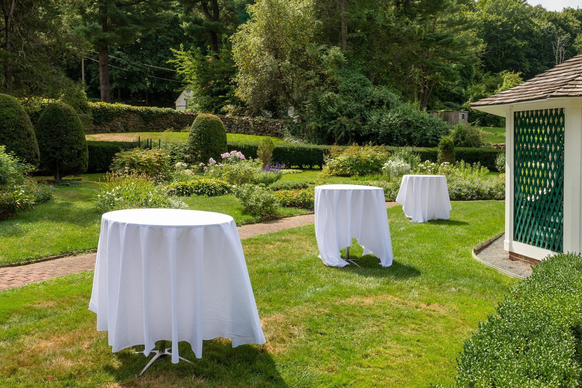 Cocktail tables with white linens on a sunny lawn in a manicured garden beside a gazebo