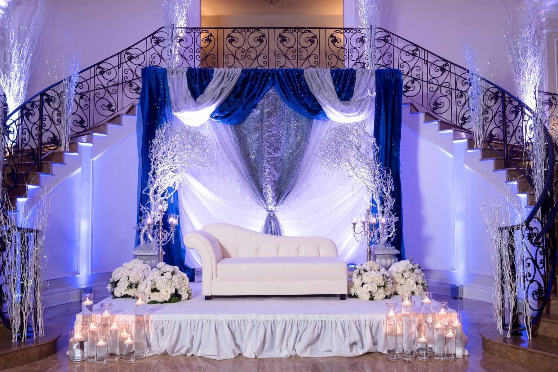 Elegant white and blue wedding stage with draped fabric, floral arrangements, and a couch under a curved staircase