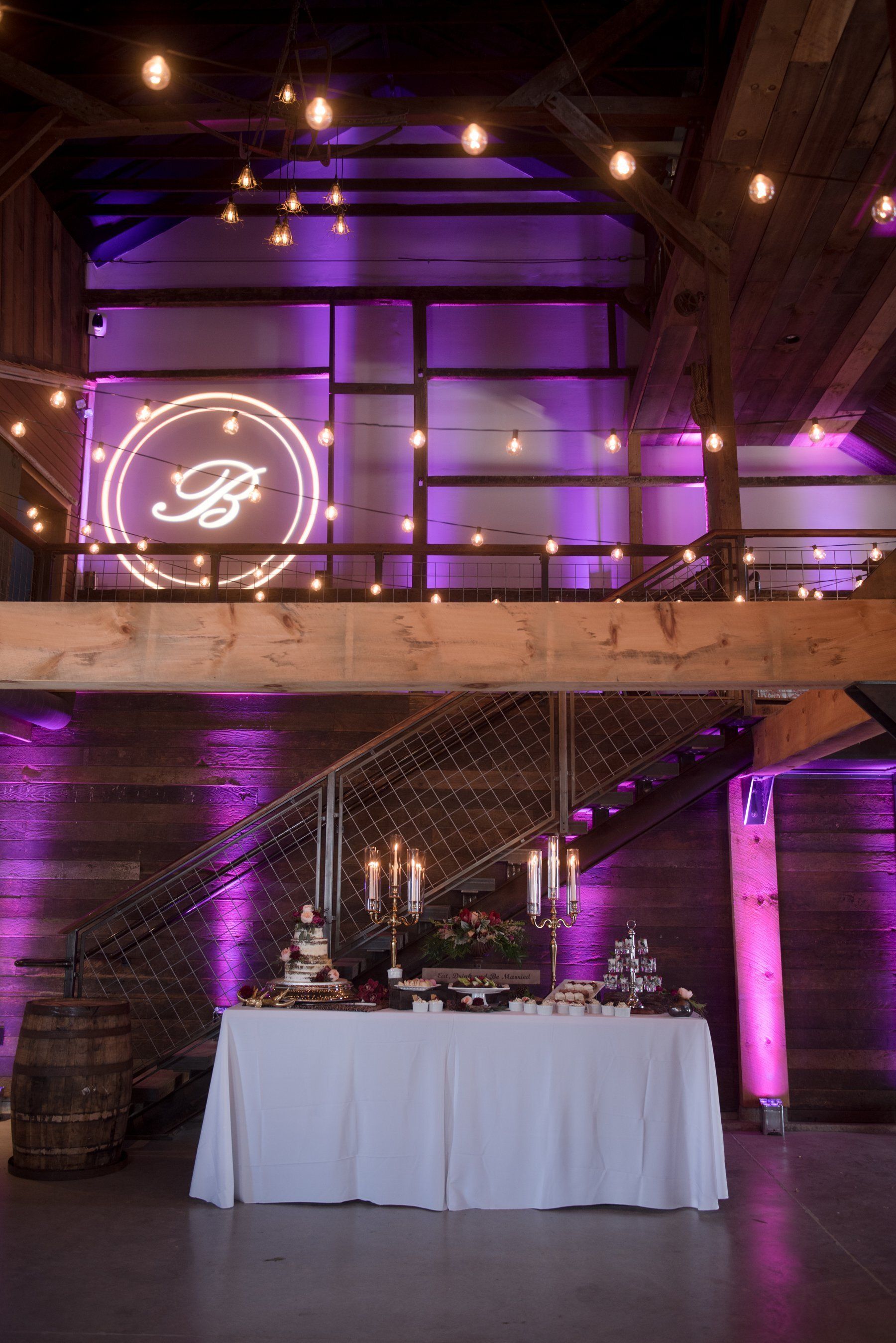 Purple-lit banquet hall with a decorated table beneath a balcony and glowing monogram sign