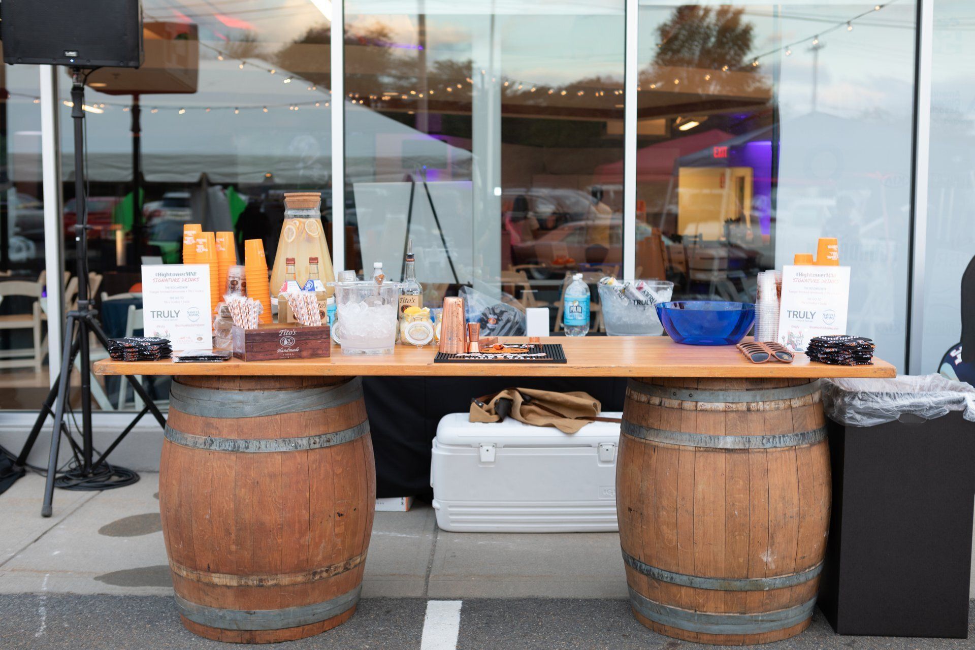 Outdoor vendor table with bottles, jars, and a cooler between two wooden barrels in front of a glass building
