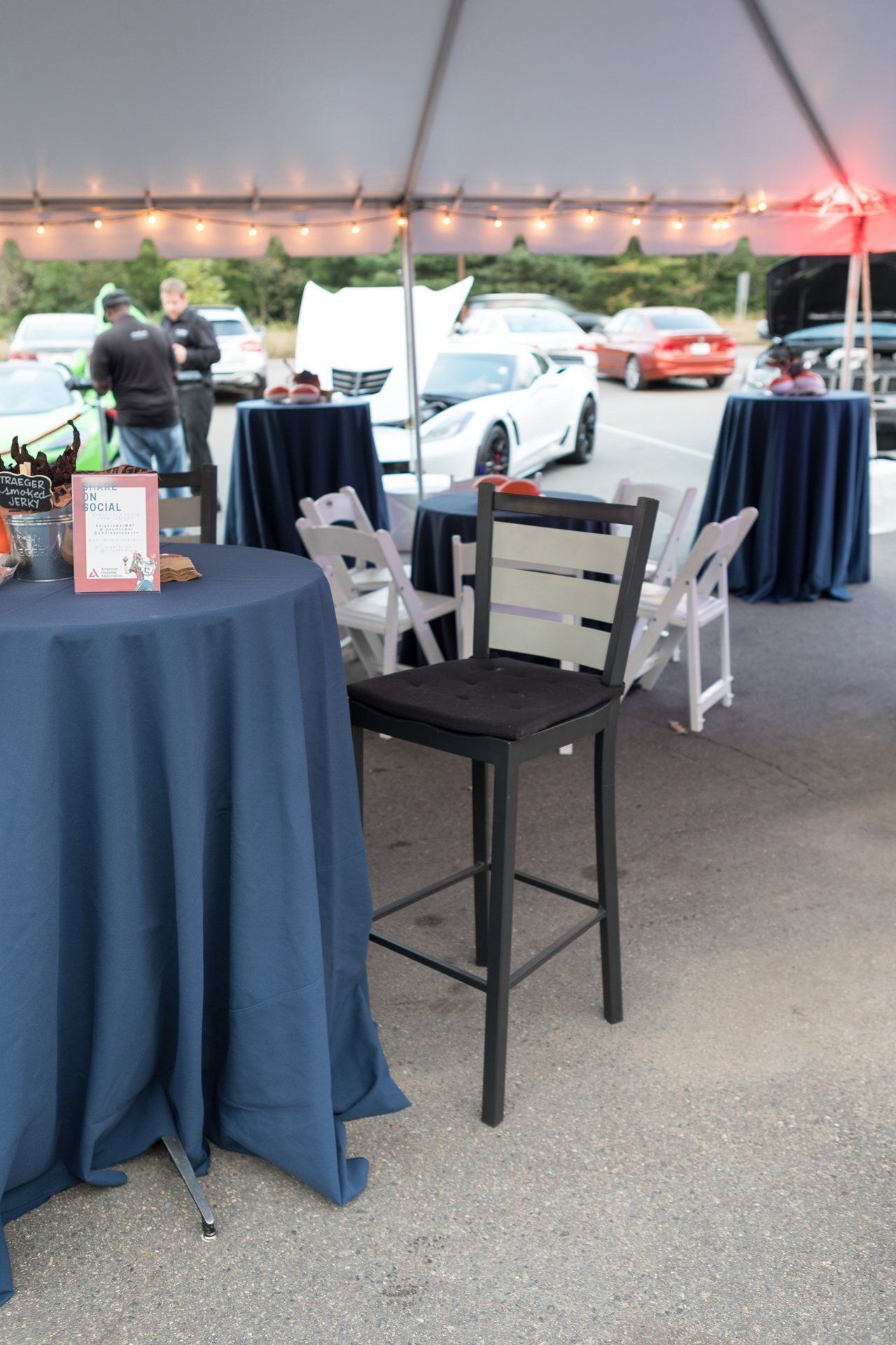 Outdoor event tent with cocktail tables, chairs, and classic cars parked in the background.