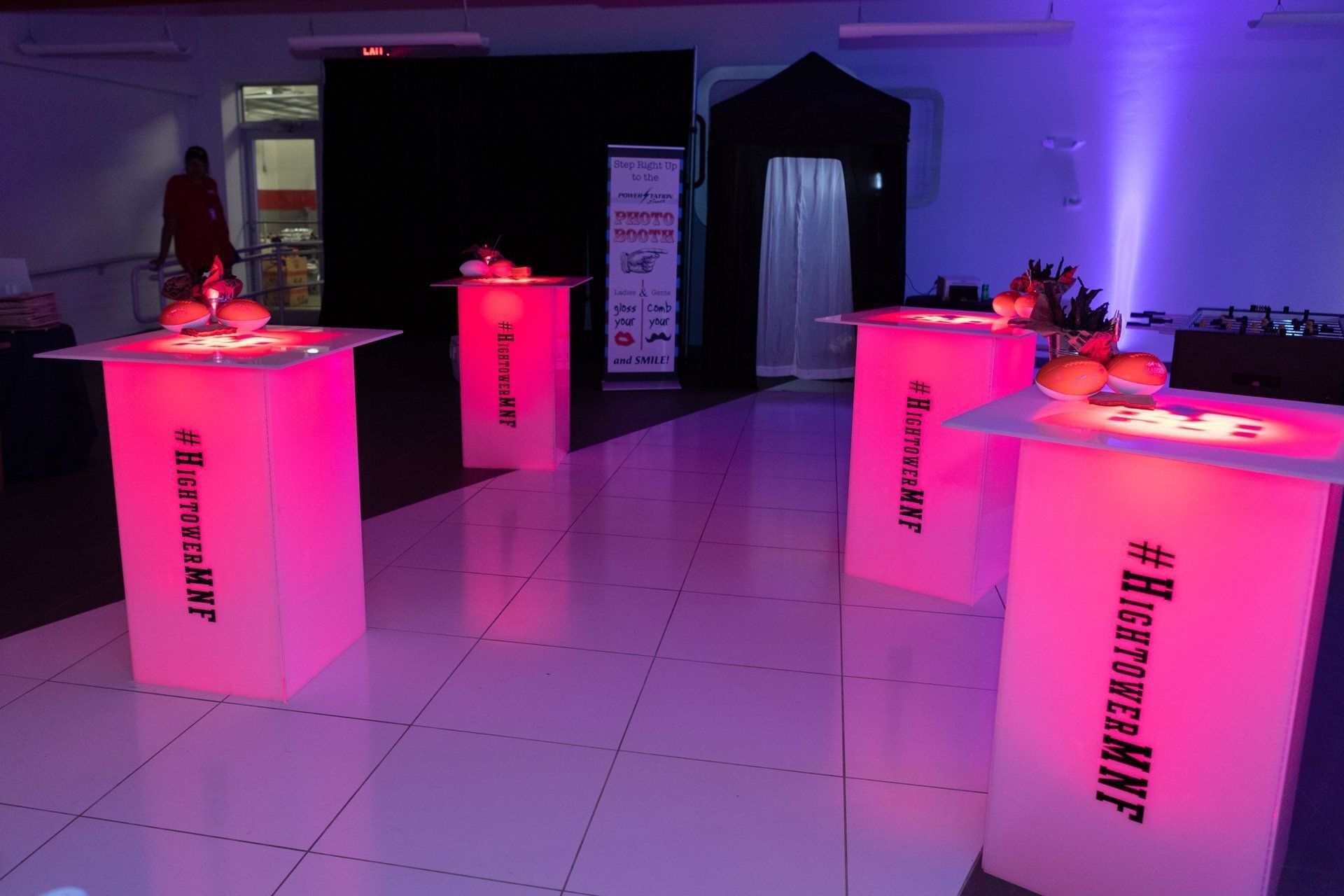 Purple-lit event room with glowing pink cocktail tables and a black photo booth in back