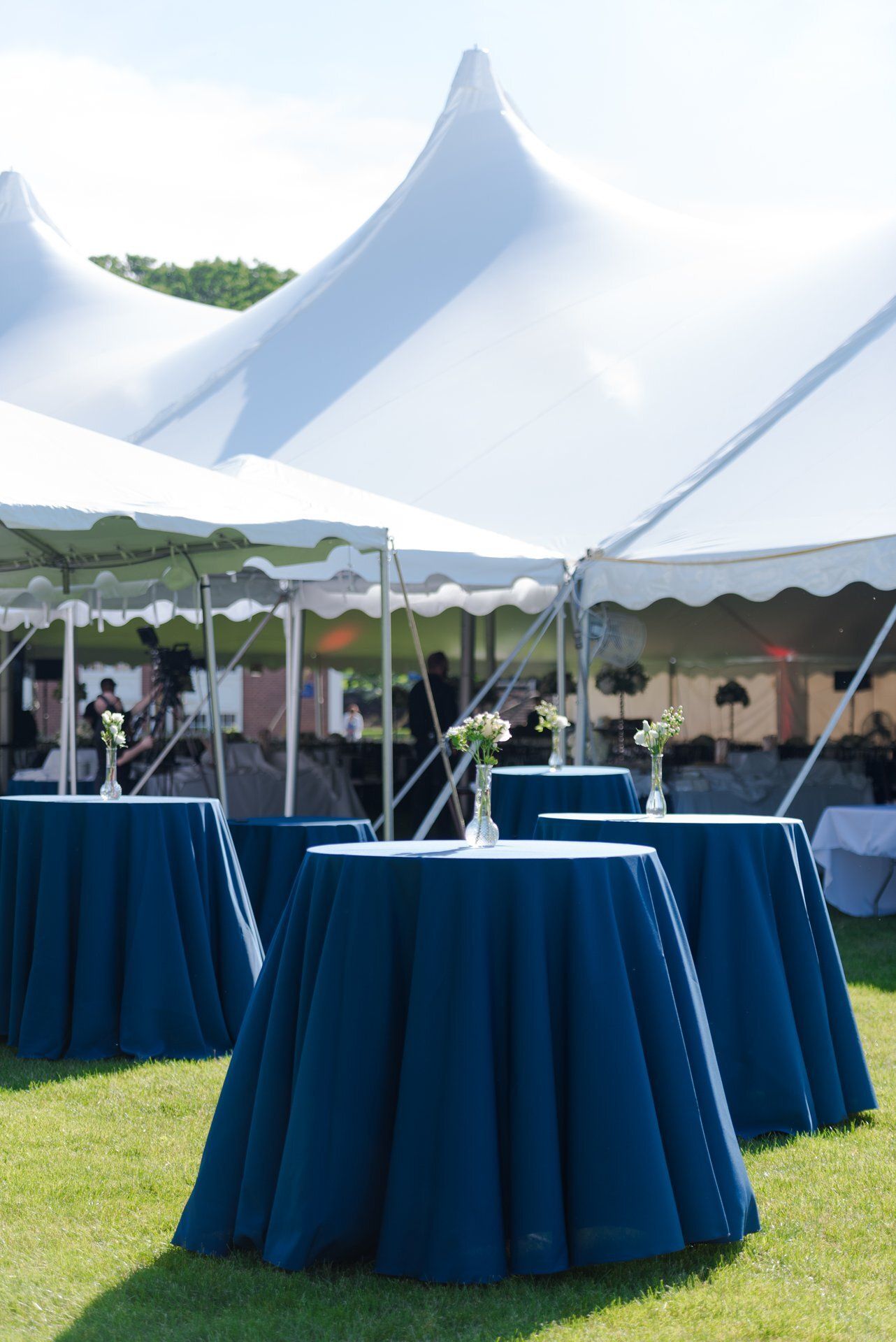 Blue-covered cocktail tables under a white event tent on a grassy lawn.