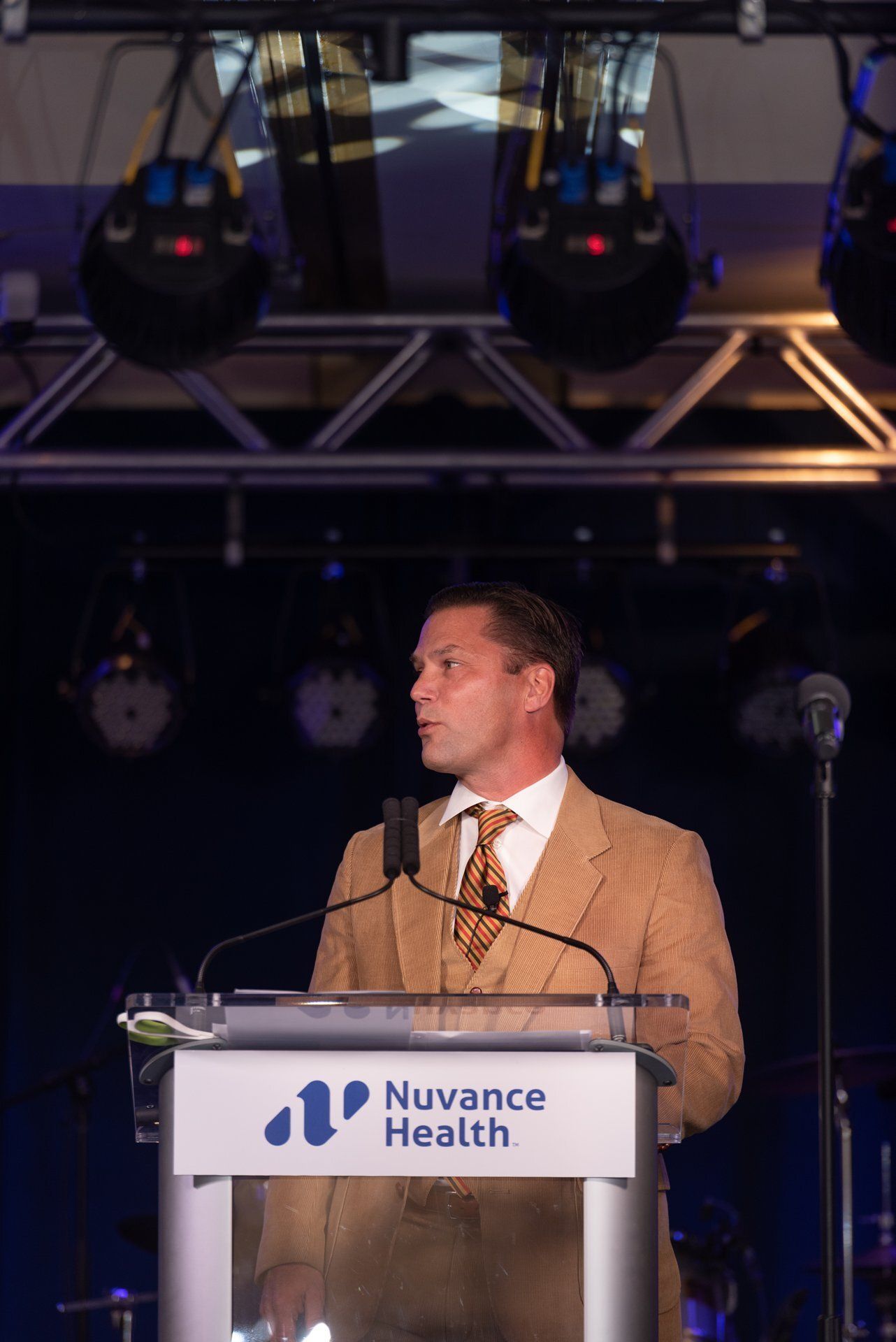 Speaker at a podium with Nuvance Health logo, looking left under stage lights.