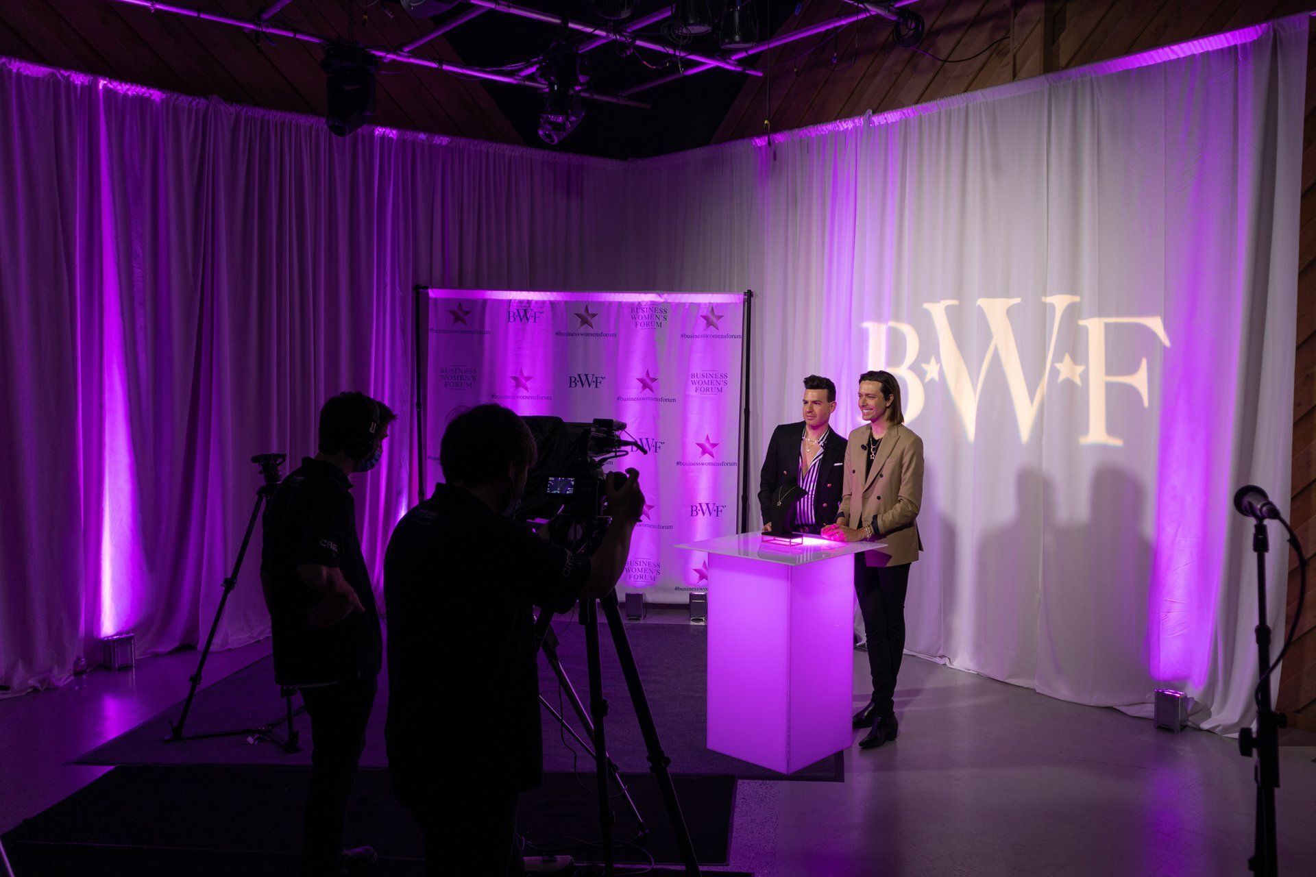 Purple-lit event stage with two people speaking at a podium, cameras filming in the foreground