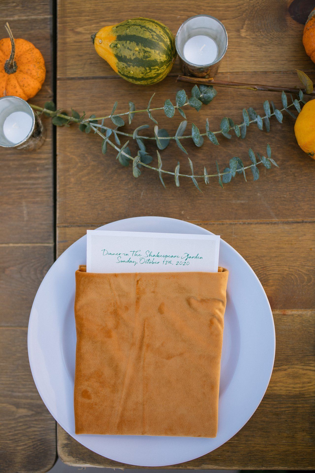 White plate with an orange napkin and card on a wooden table with pumpkins and greenery