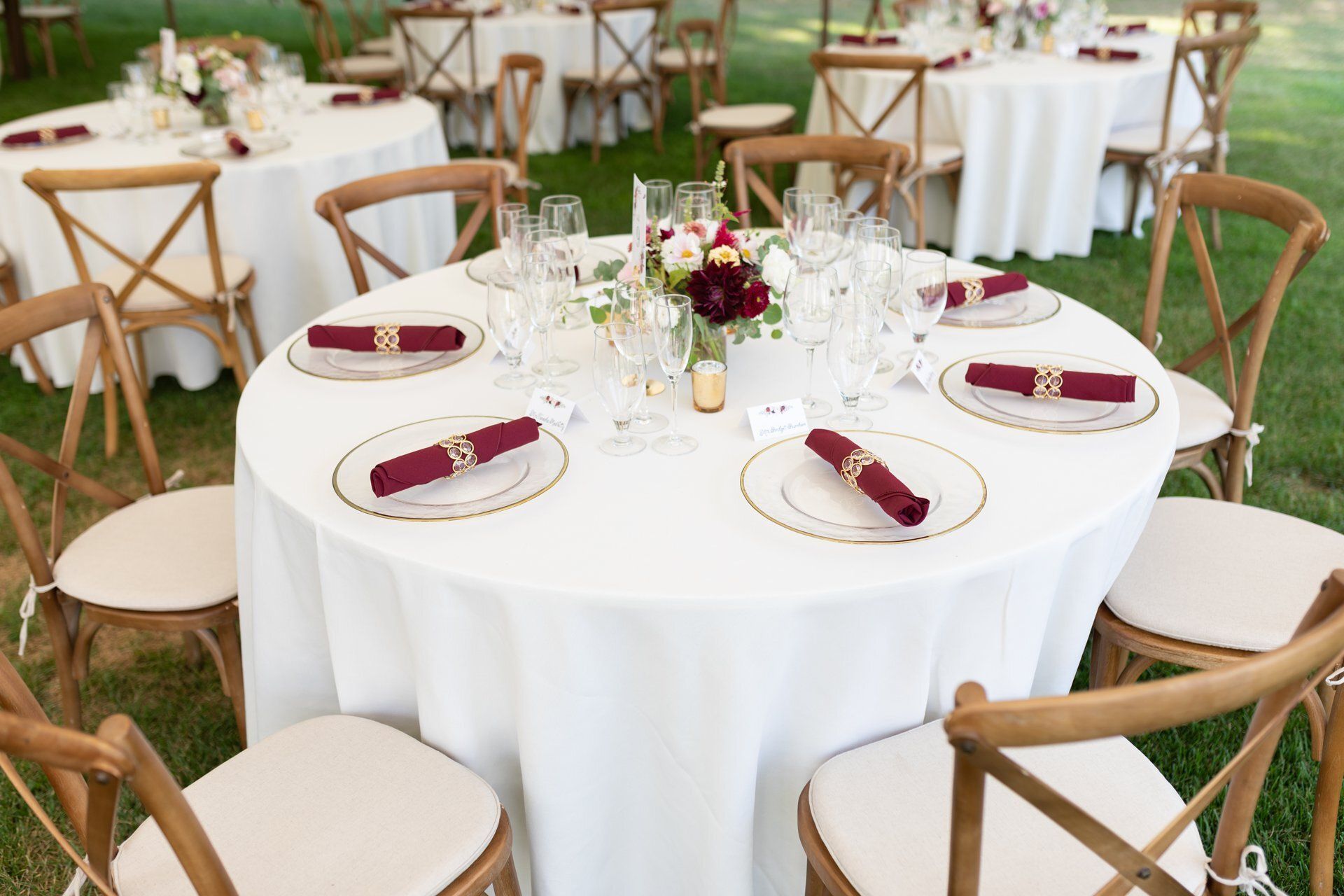 Round banquet tables with white linens and burgundy napkins set on woven chairs outdoors on grass