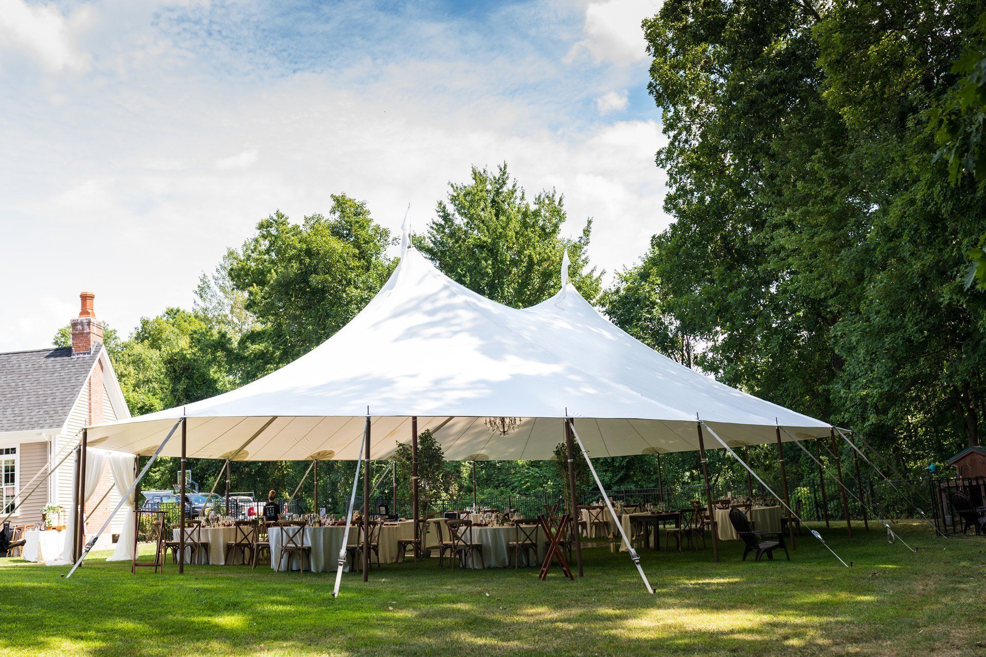 White event tent set up on a grassy lawn with tables underneath, surrounded by trees on a sunny day