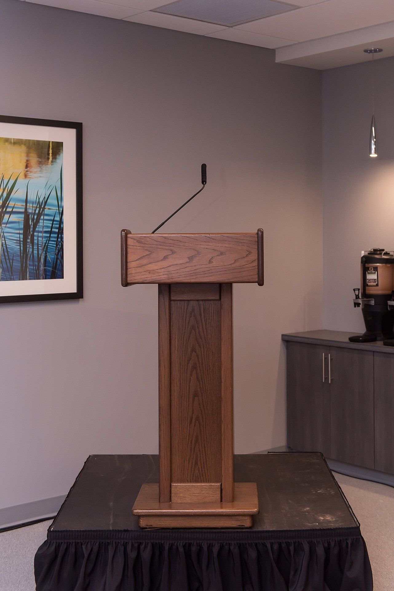 Wooden podium with microphone in a conference room, on a stage beside a framed wall print.