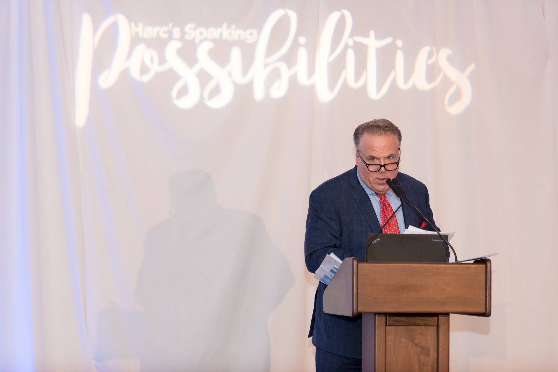 Speaker at podium before “Possibilities” backdrop, reading notes during a formal event