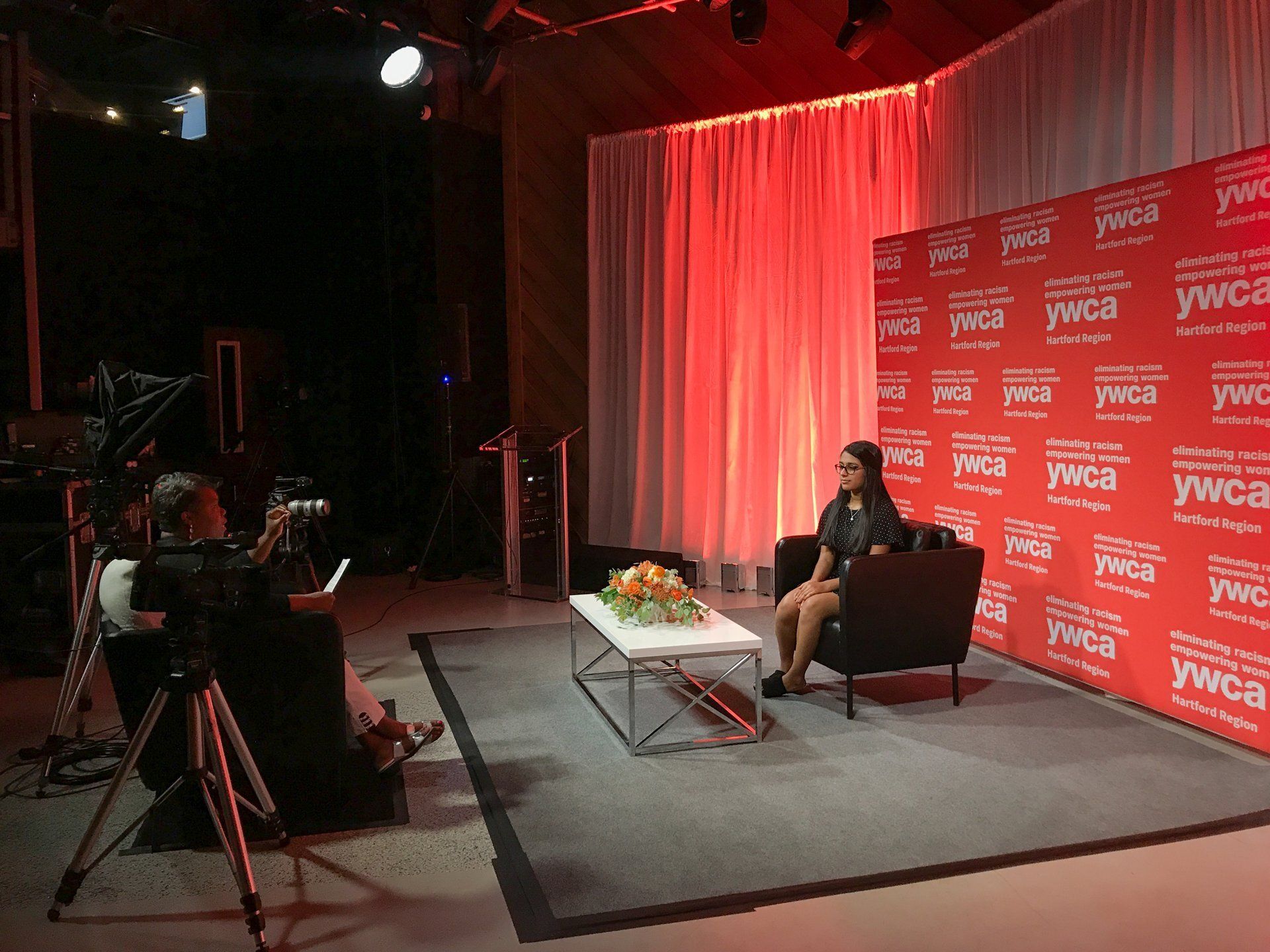 Studio interview setup with a seated guest, cameras, and red branded backdrop