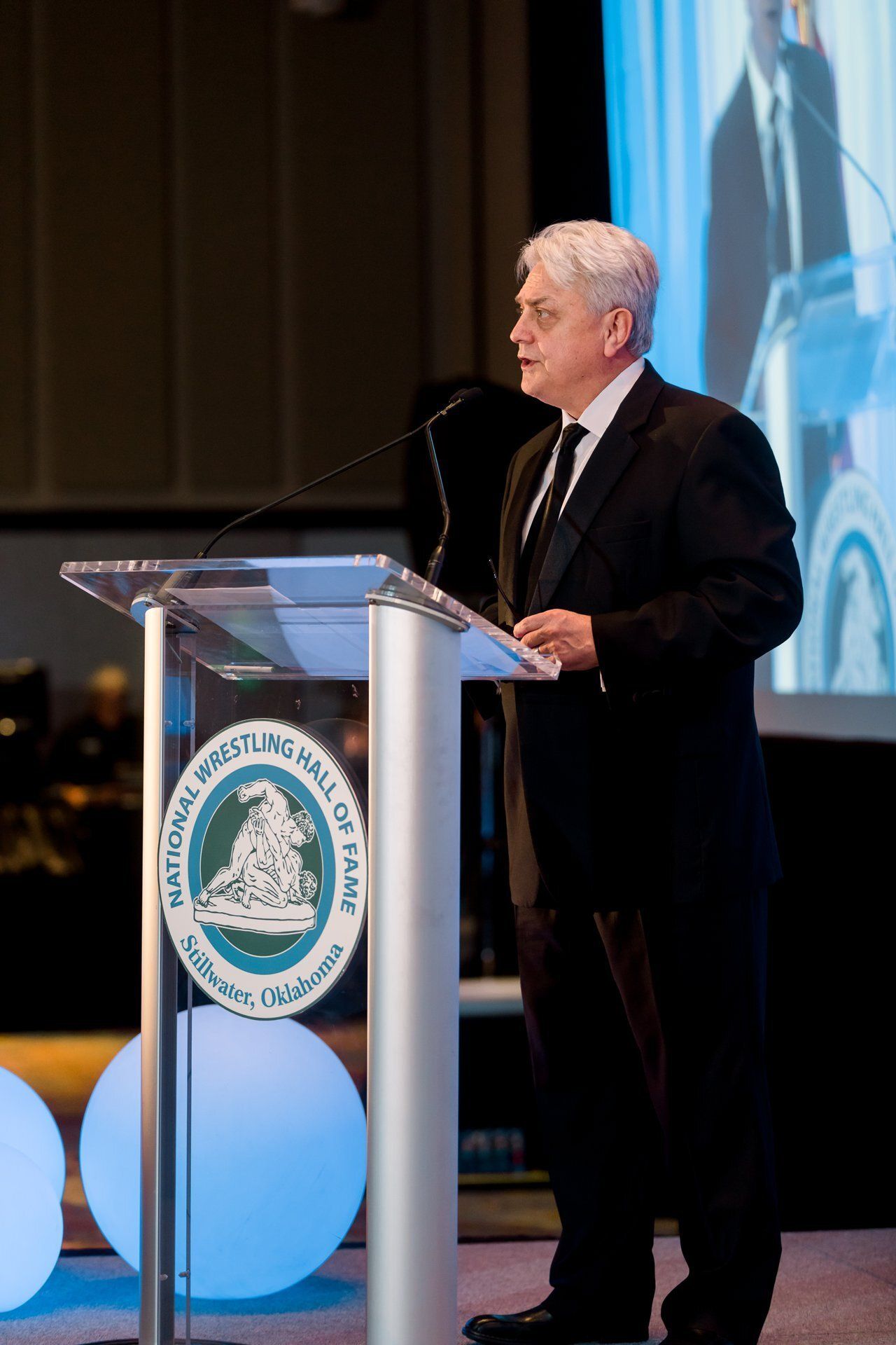 Man speaking at a podium with a seal, beside a large screen at a formal event
