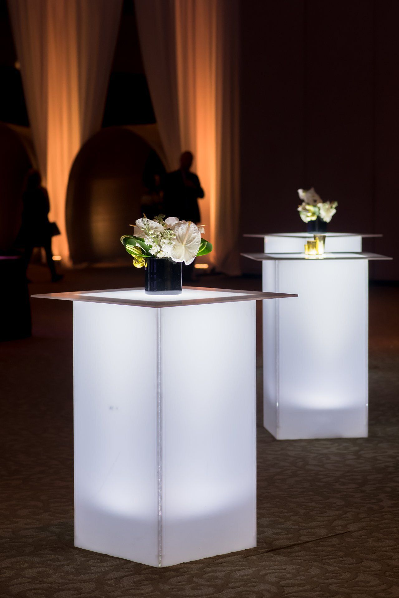 Two glowing white pedestal tables with floral arrangements in a dimly lit event space