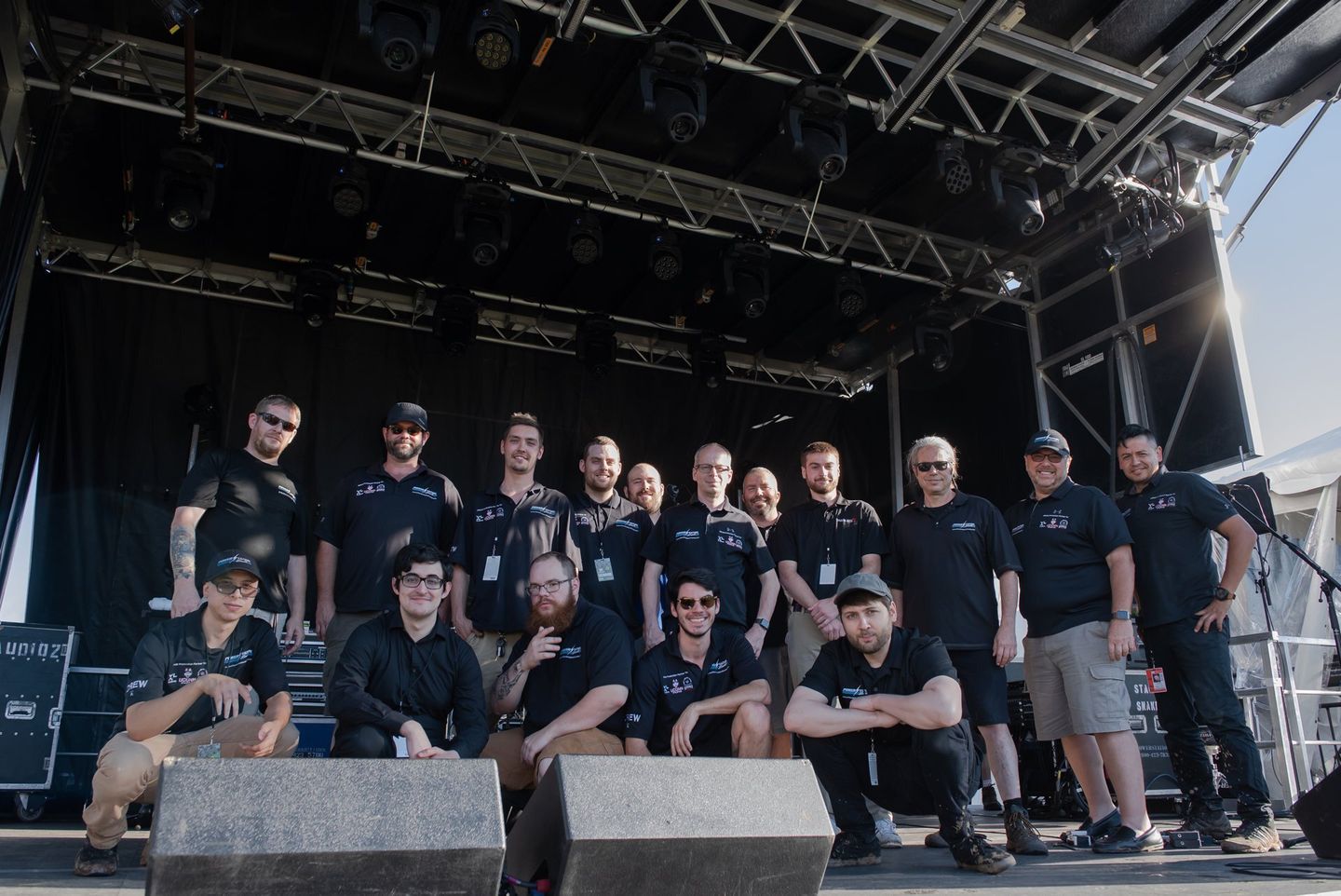 Group of people in black shirts posing on an outdoor stage under scaffolding at a concert venue